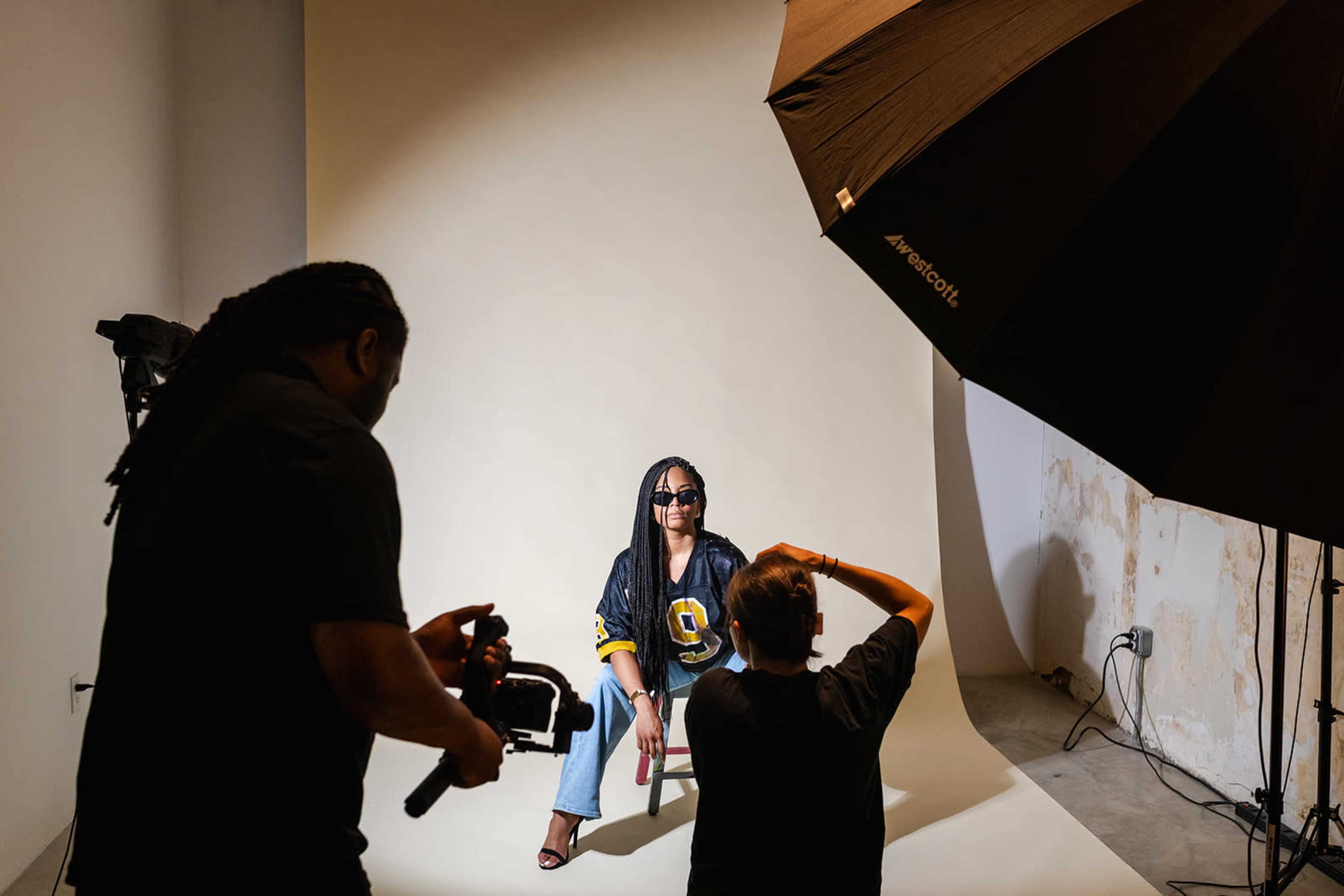 A model sits in a chair under studio lighting while two photographers adjust their equipment in a photography studio.