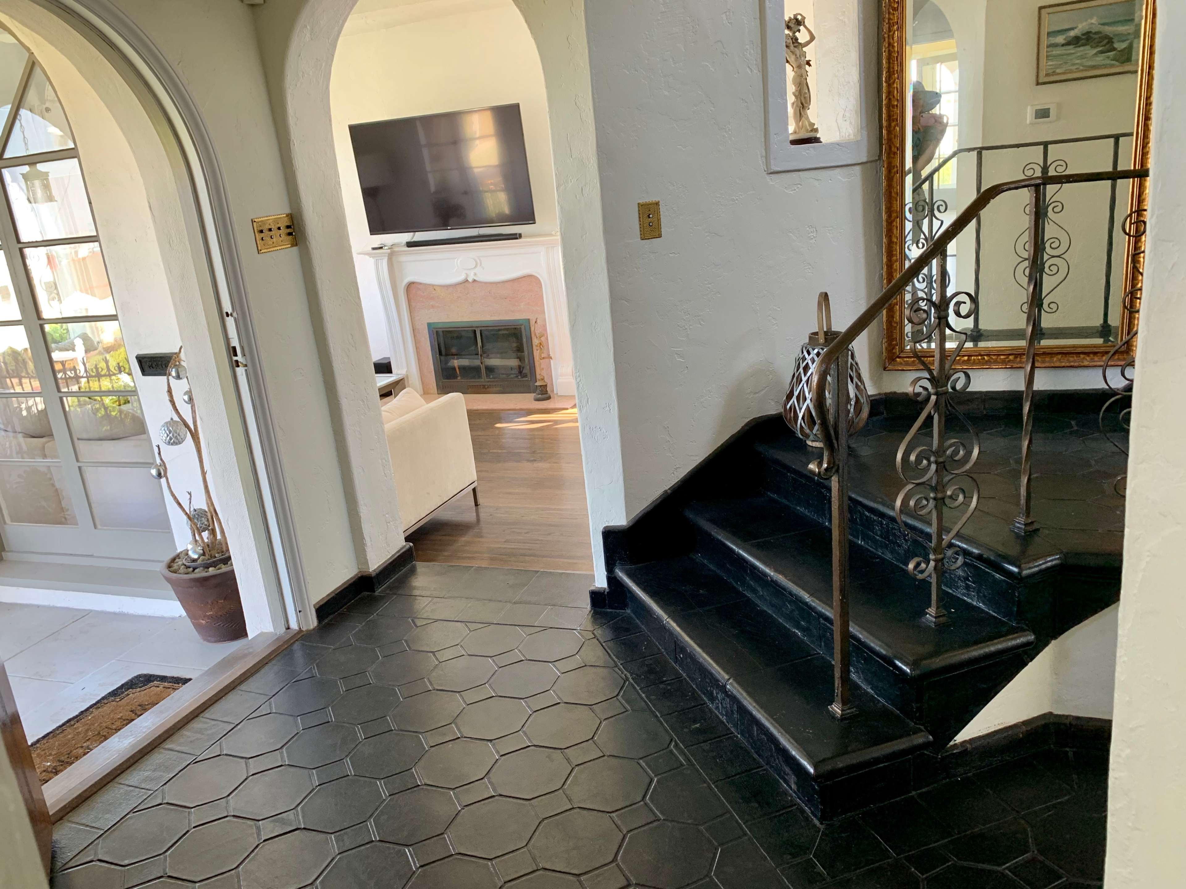 A hallway with a staircase leading to an upper level, a living room with a fireplace visible in the background, and decorative elements like a mirror and a plant.
