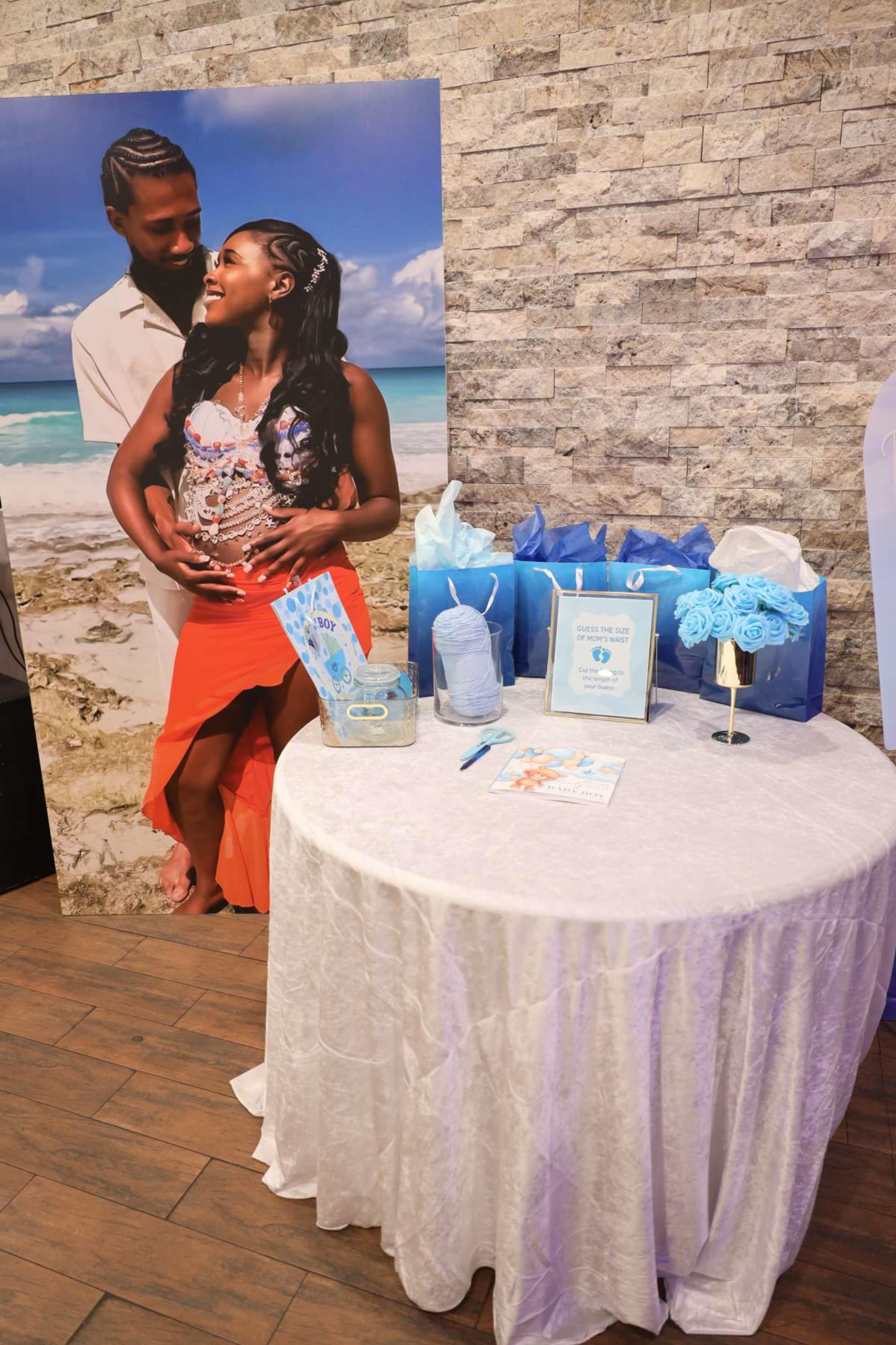 A table adorned with a light-colored cloth displays blue gift bags, a framed picture, and a guest book, set against a backdrop featuring a couple by the beach.