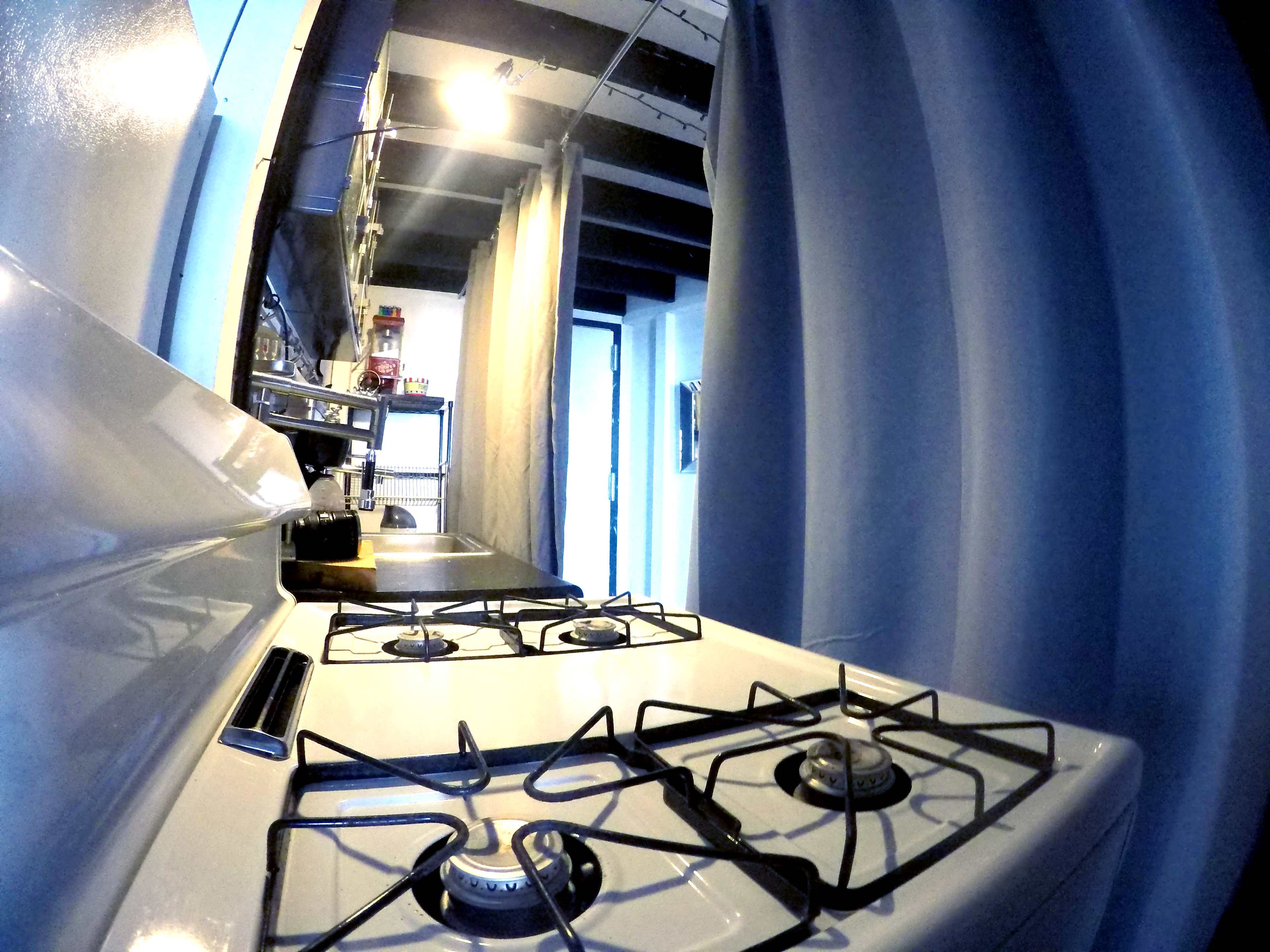 The image shows a kitchen with a white gas stove on the left and a doorway leading to another room at the end of a narrow hallway, flanked by blue curtains.