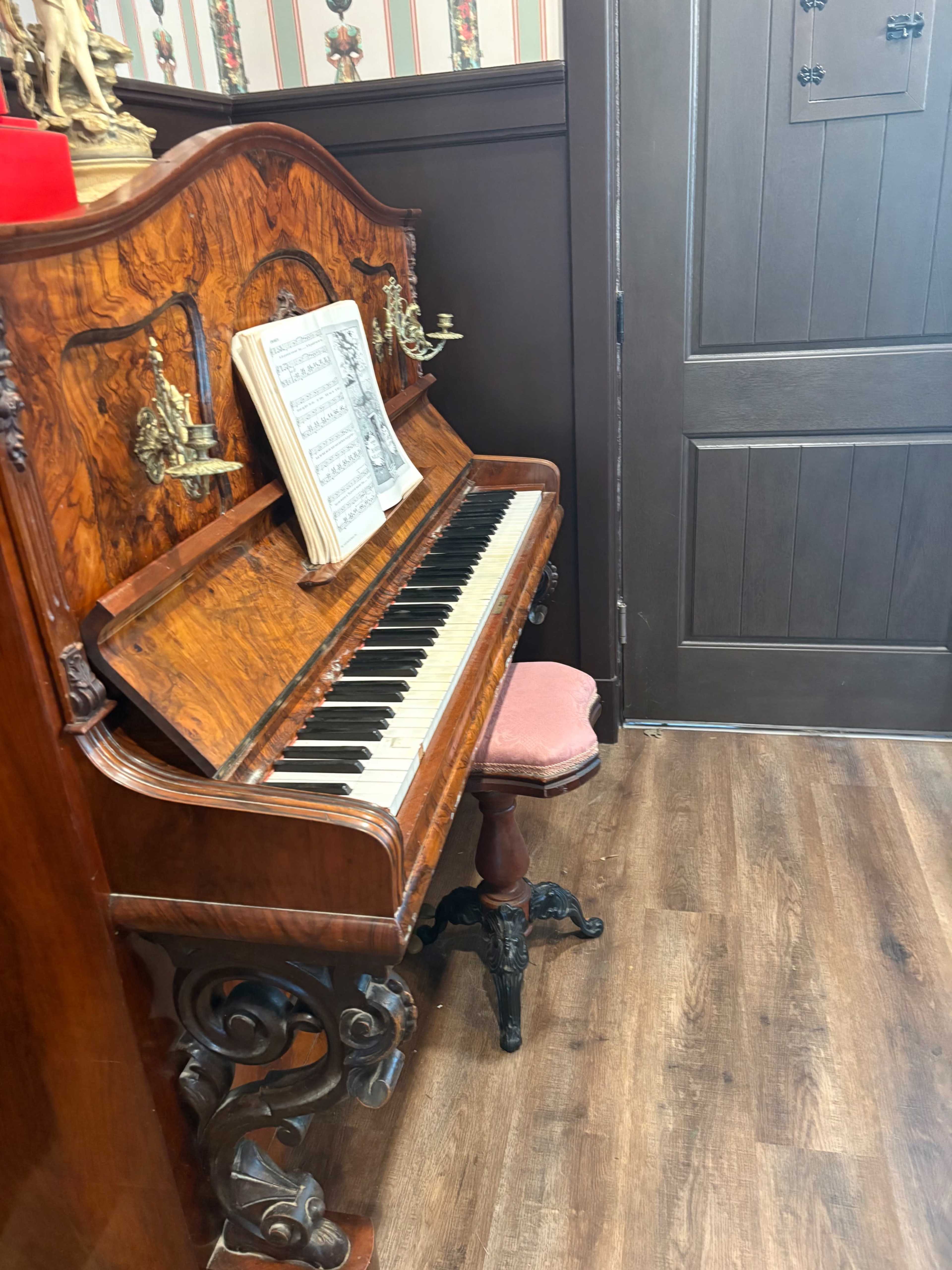 A vintage wooden piano with ornate details sits against a wall, accompanied by a music sheet on its stand and a small upholstered stool.