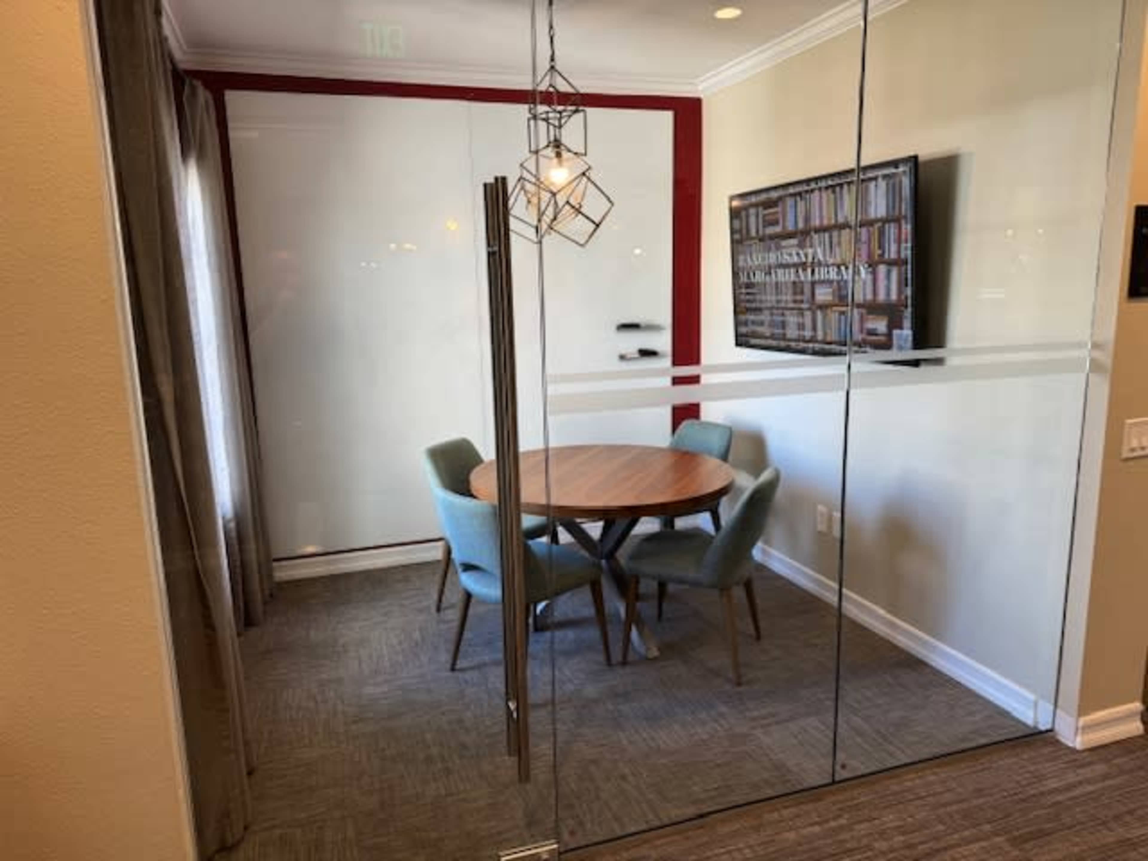 A glass-walled meeting room features a round wooden table surrounded by four light blue chairs and a wall-mounted TV displaying a bookshelf graphic.