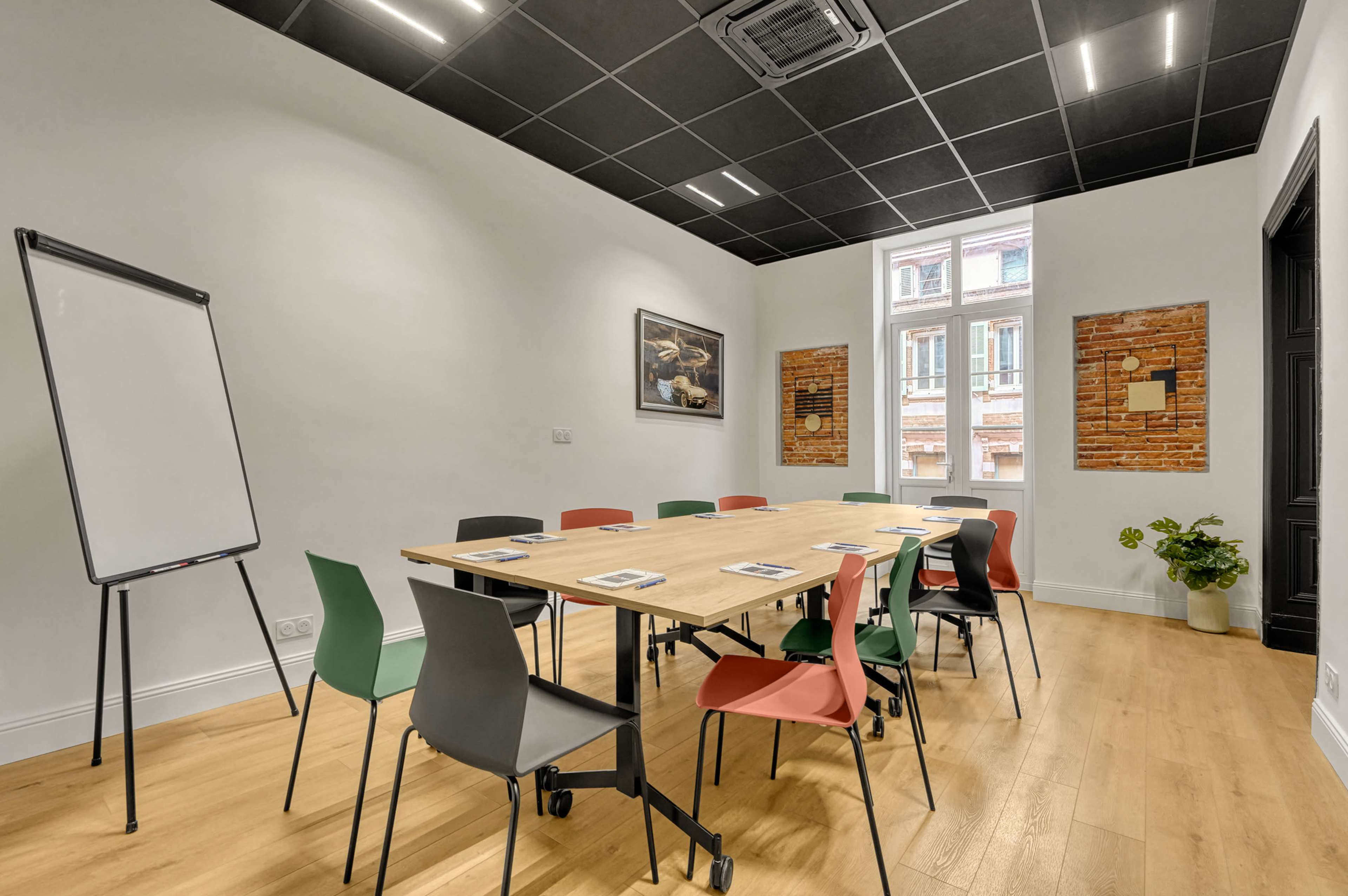 A modern conference room features a large wooden table surrounded by colorful chairs, a whiteboard, and large windows with an exposed brick wall backdrop.