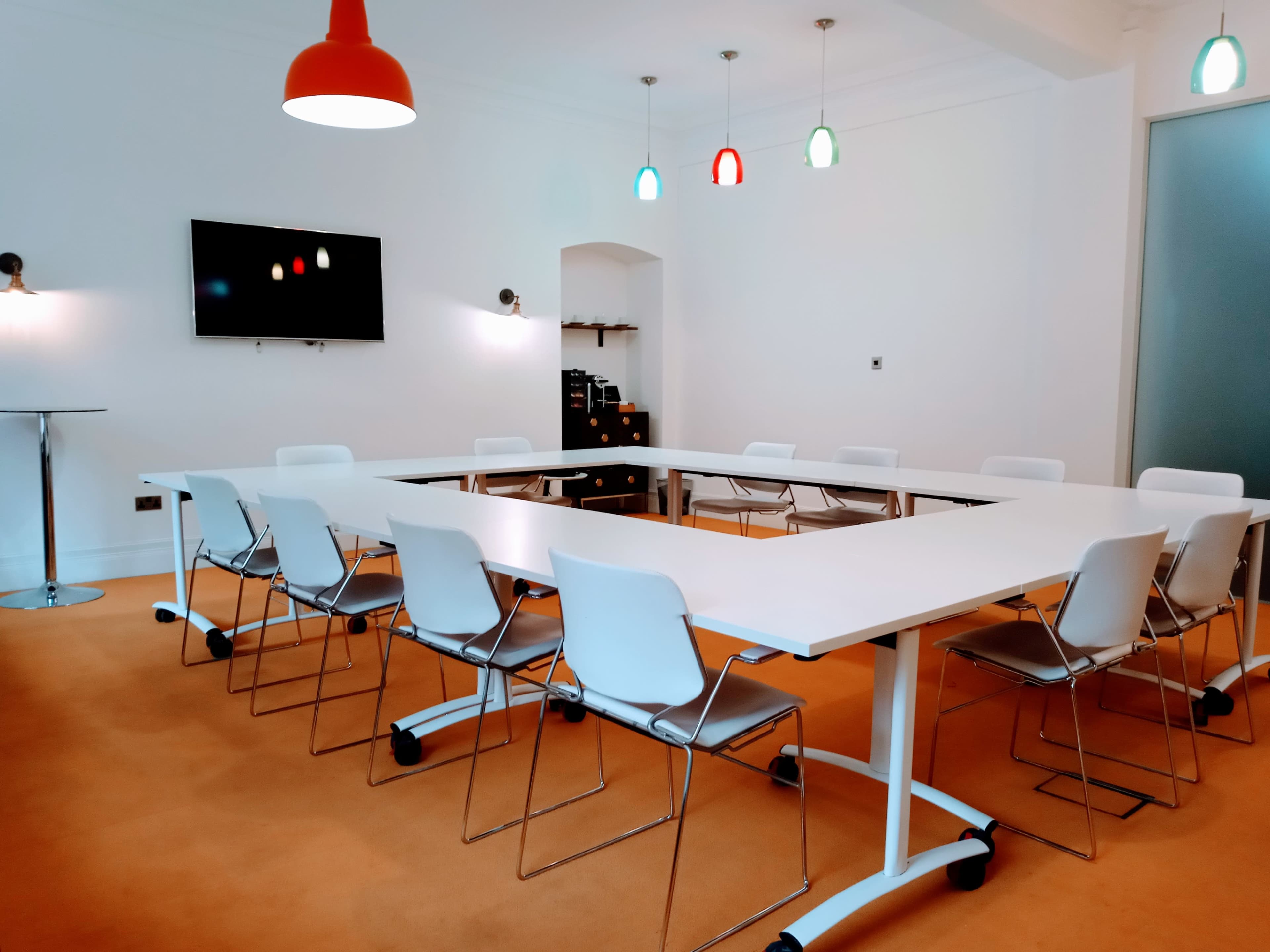 A modern conference room features a large white table surrounded by eight white chairs, with a television mounted on the wall and colorful pendant lights hanging from the ceiling.