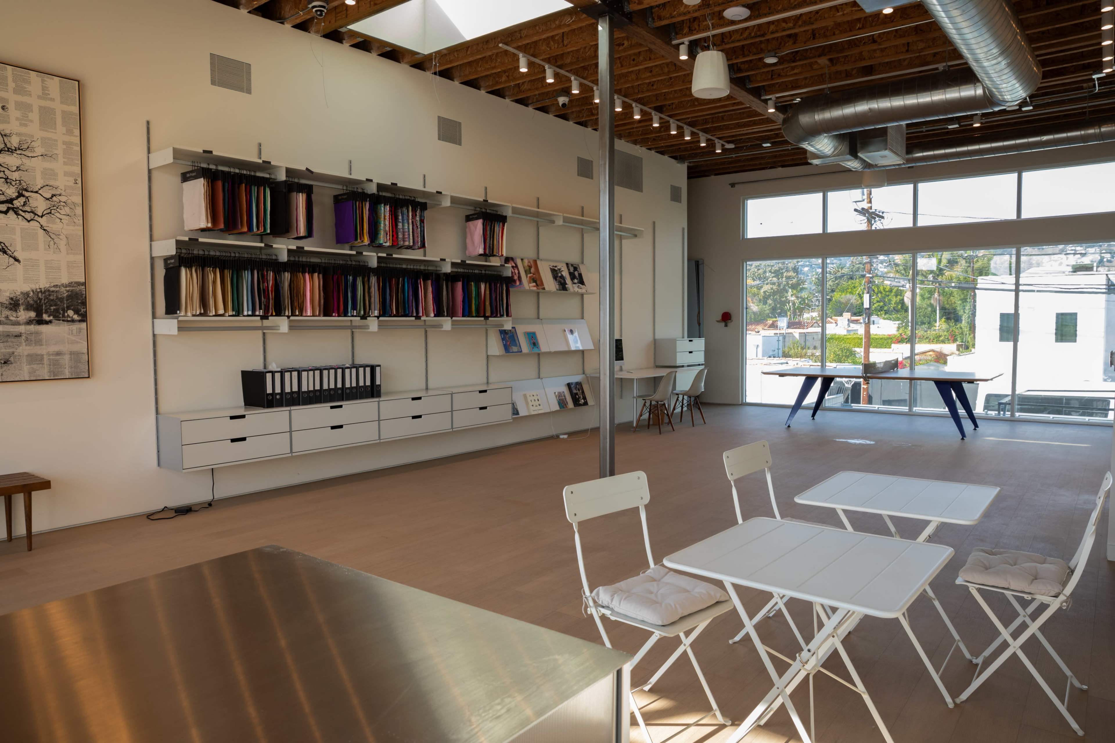 The interior of a modern workspace features a minimalist design with shelves of colorful fabrics, a long table, and seating arrangements.