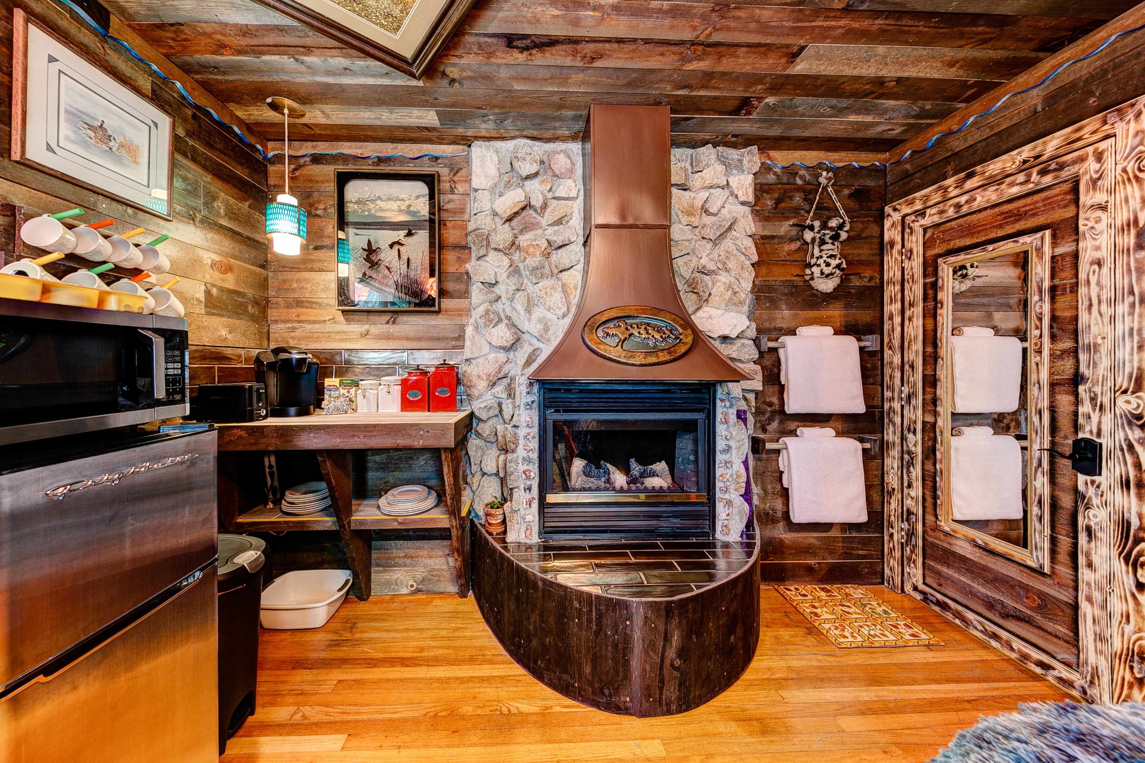 A rustic cabin interior featuring a stone fireplace adorned with a copper hood, wooden walls, and a kitchenette area.