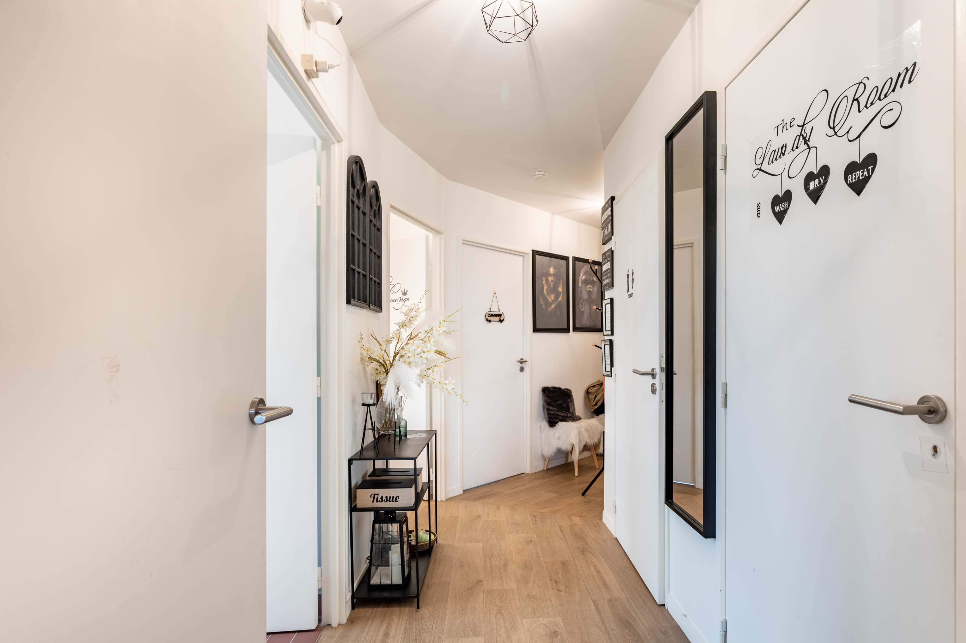 A hallway with white walls, wooden flooring, and multiple doors leading to different rooms, along with decorative elements like a plant and framed pictures.