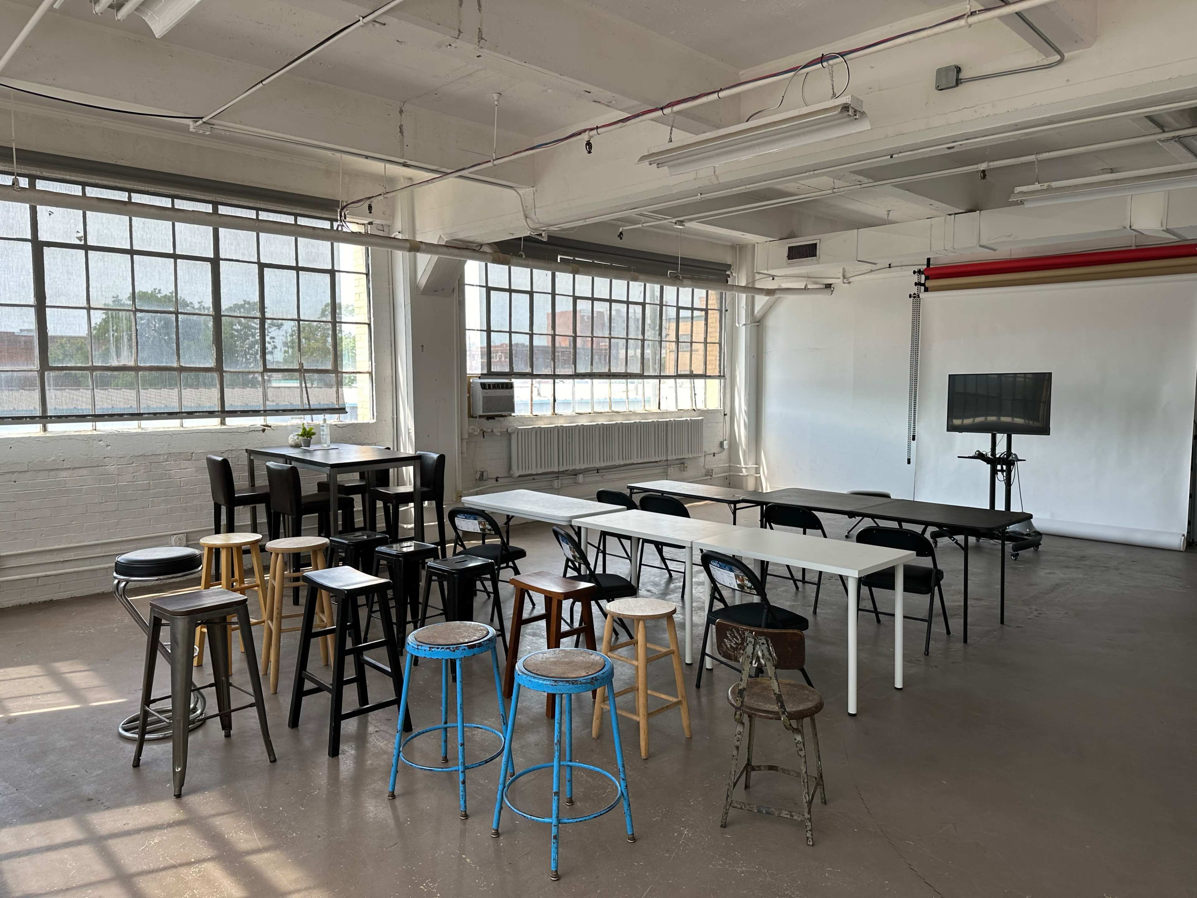 The image shows a spacious, bright room with large windows, furnished with various types of stools and tables arranged in different configurations.