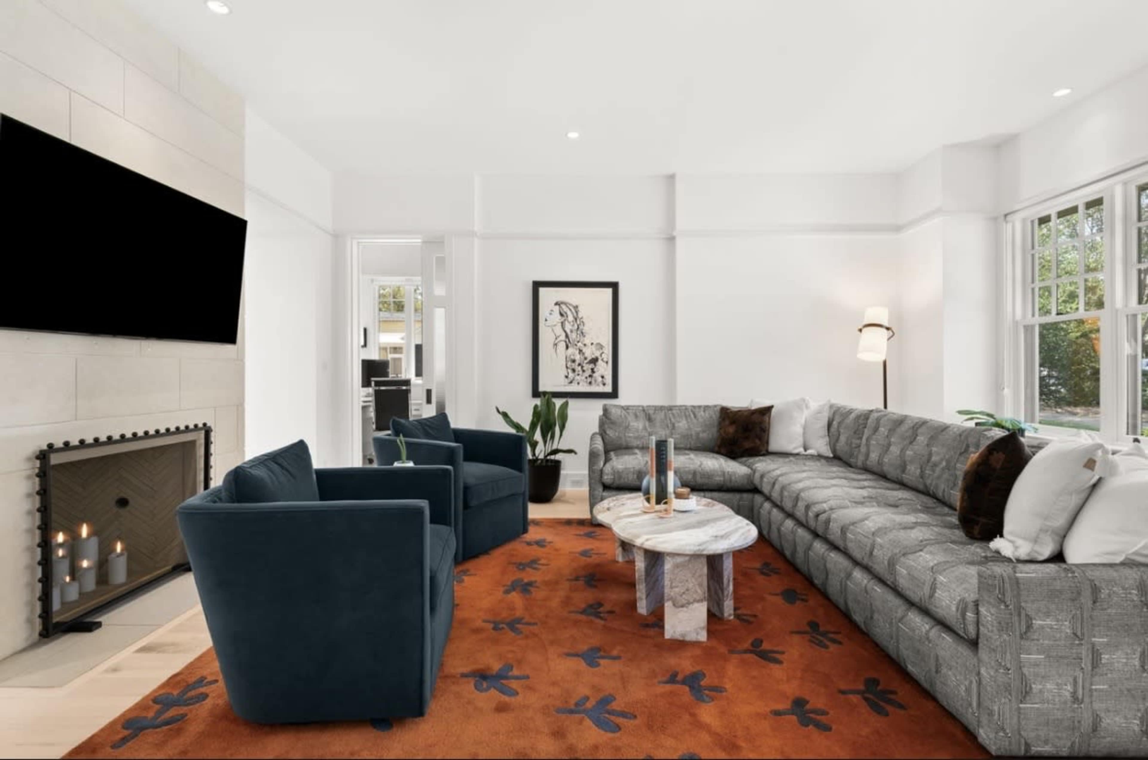 The image shows a modern living room with a large gray sectional sofa, two blue armchairs, a marble coffee table, and a fireplace, all set on an orange patterned rug.