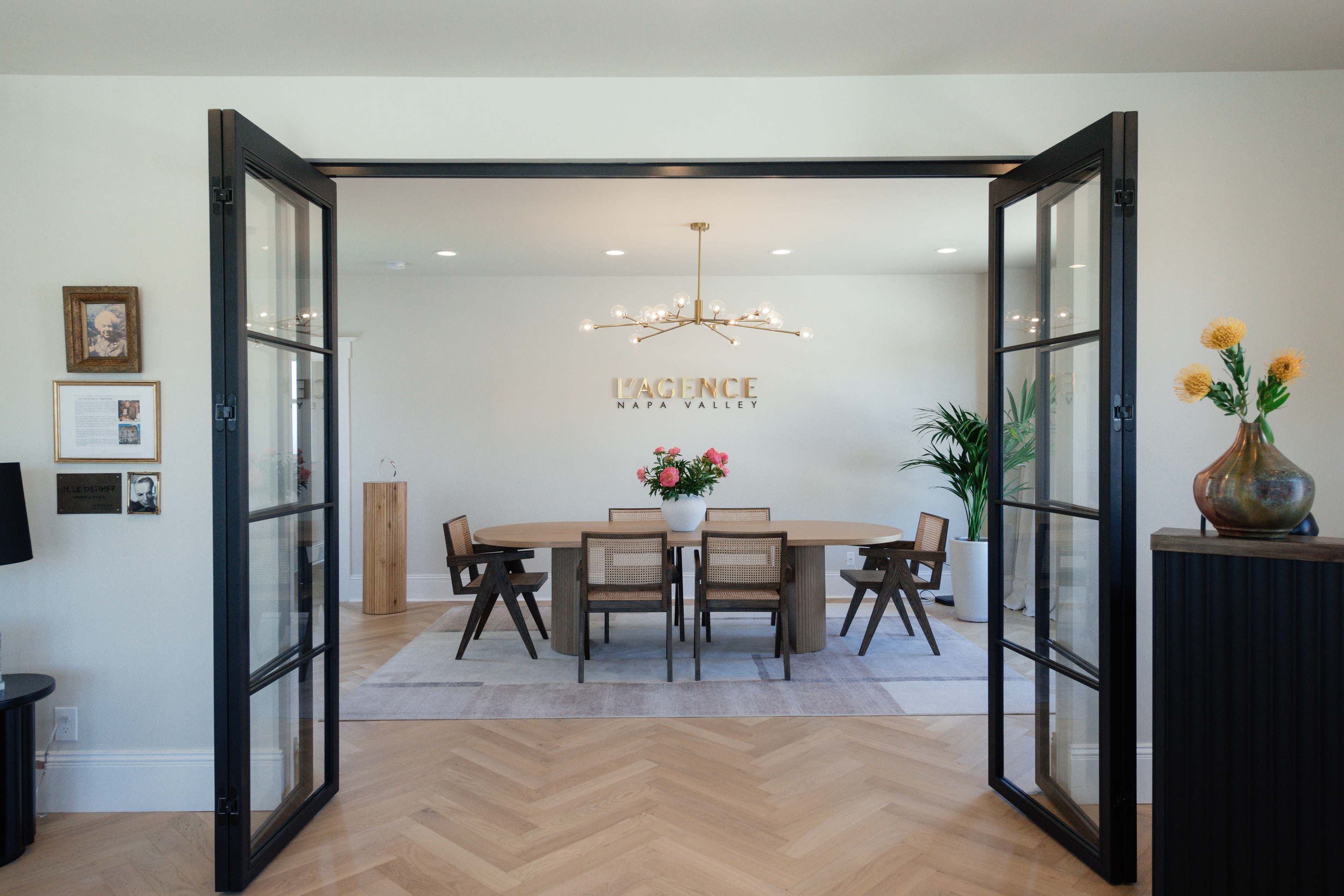 A spacious dining area with a large round table surrounded by chairs, framed by double doors and featuring a modern chandelier overhead.