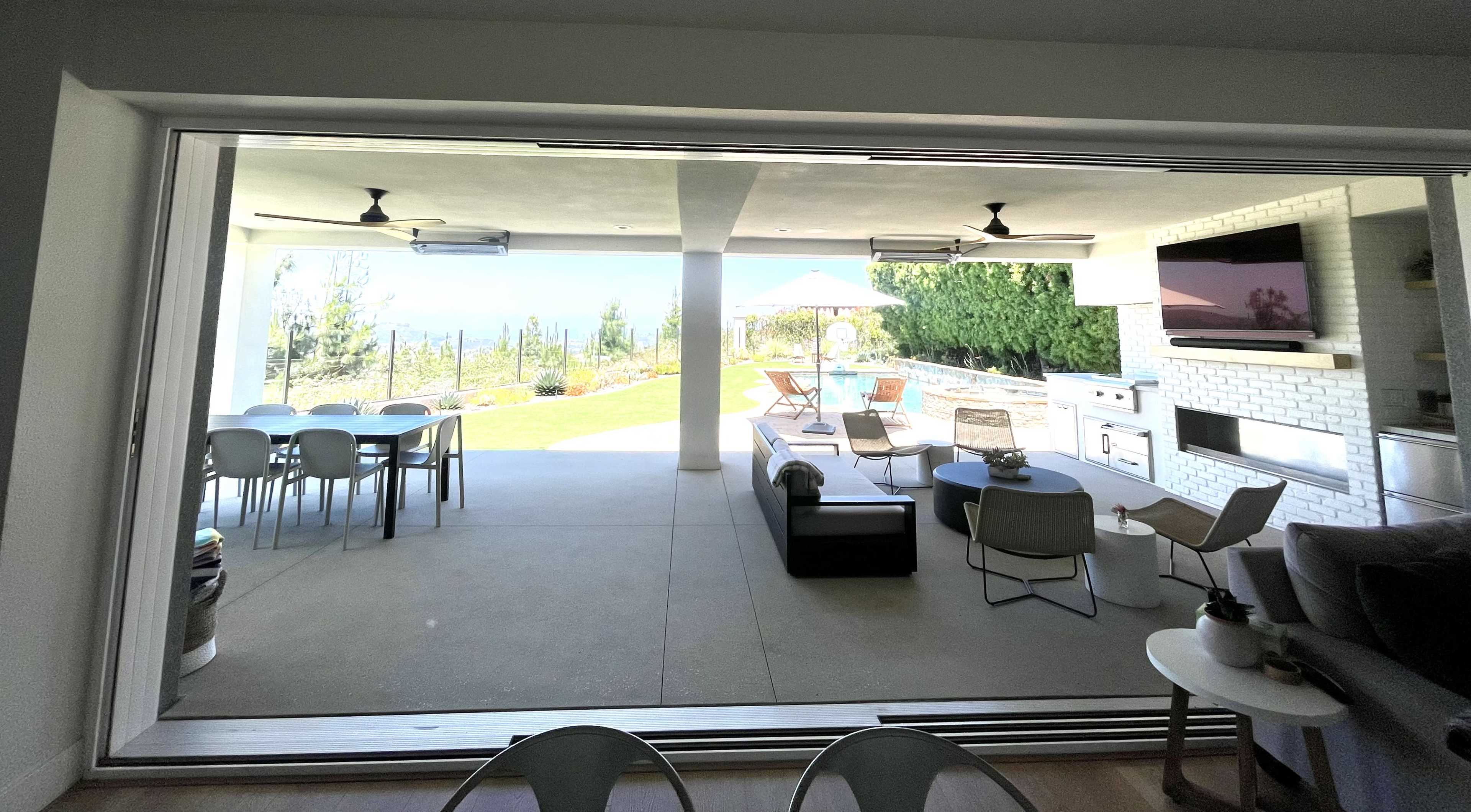 A modern indoor-outdoor living space with a view of a patio featuring dining and lounge furniture, an outdoor kitchen, and a swimming pool in the background.