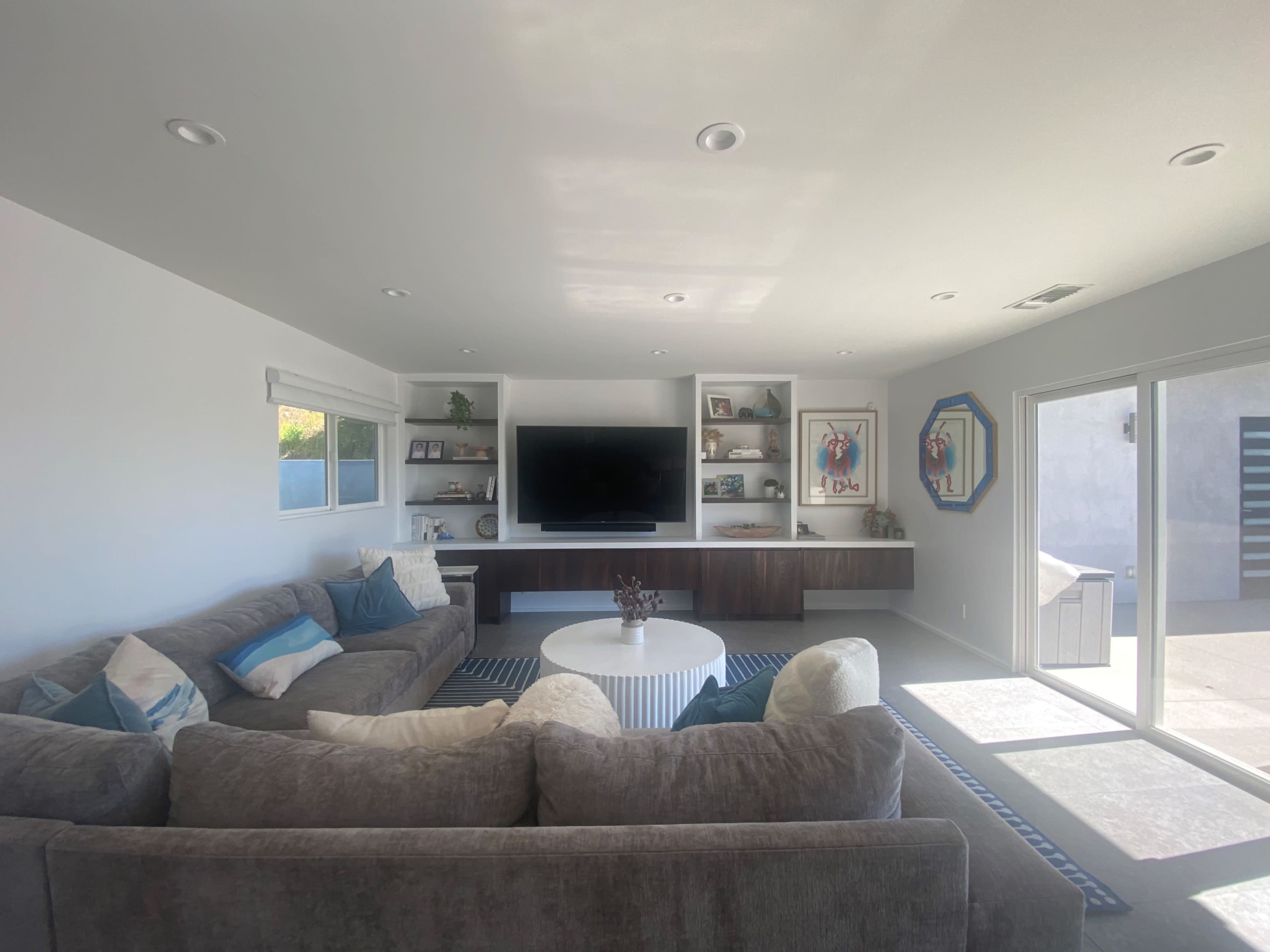 A modern living room features a large sectional sofa, a circular coffee table, and a wall-mounted TV with shelves displaying decor.