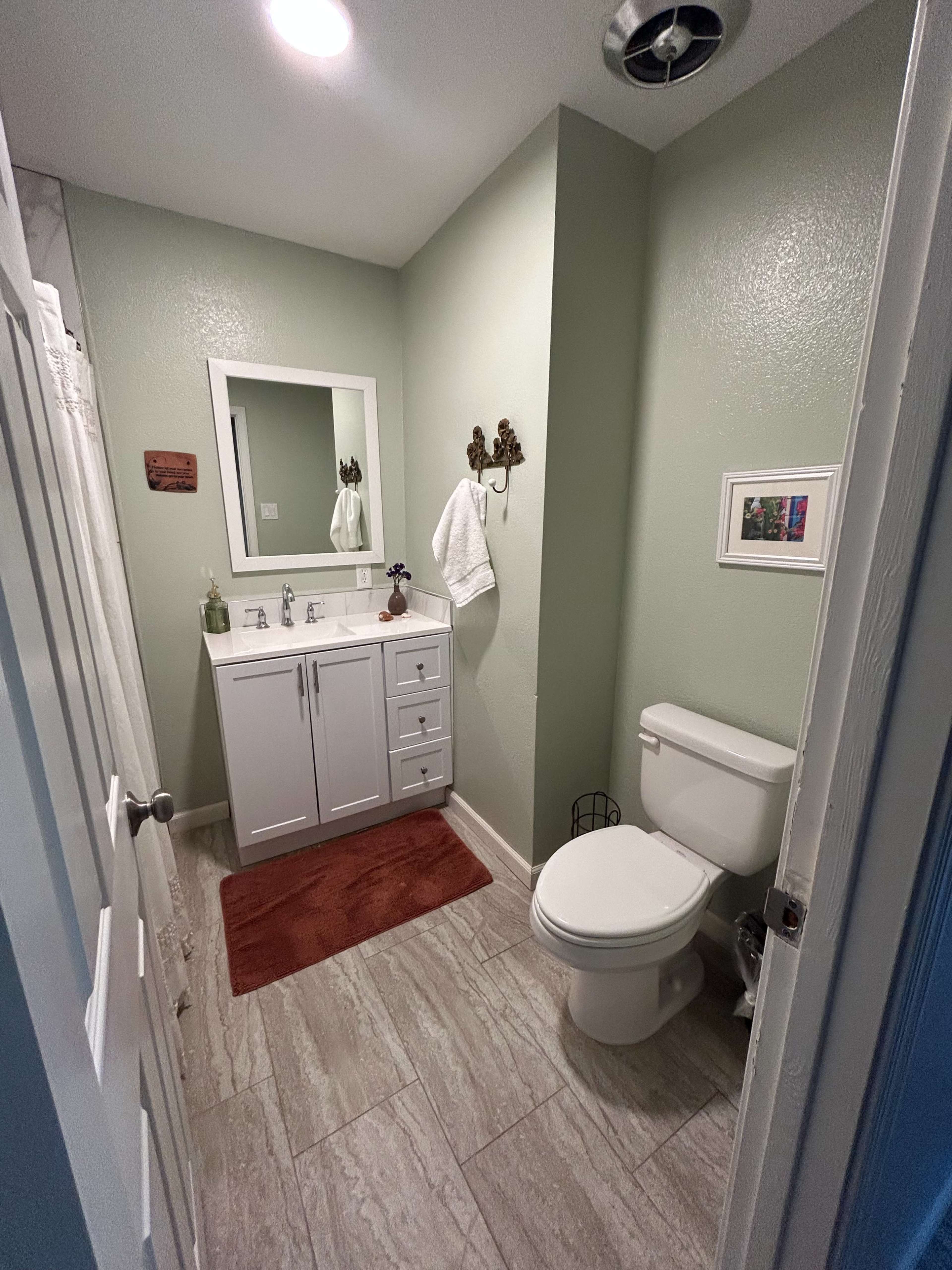 The image shows a small bathroom featuring a white vanity with a mirror, a toilet, and light green walls.