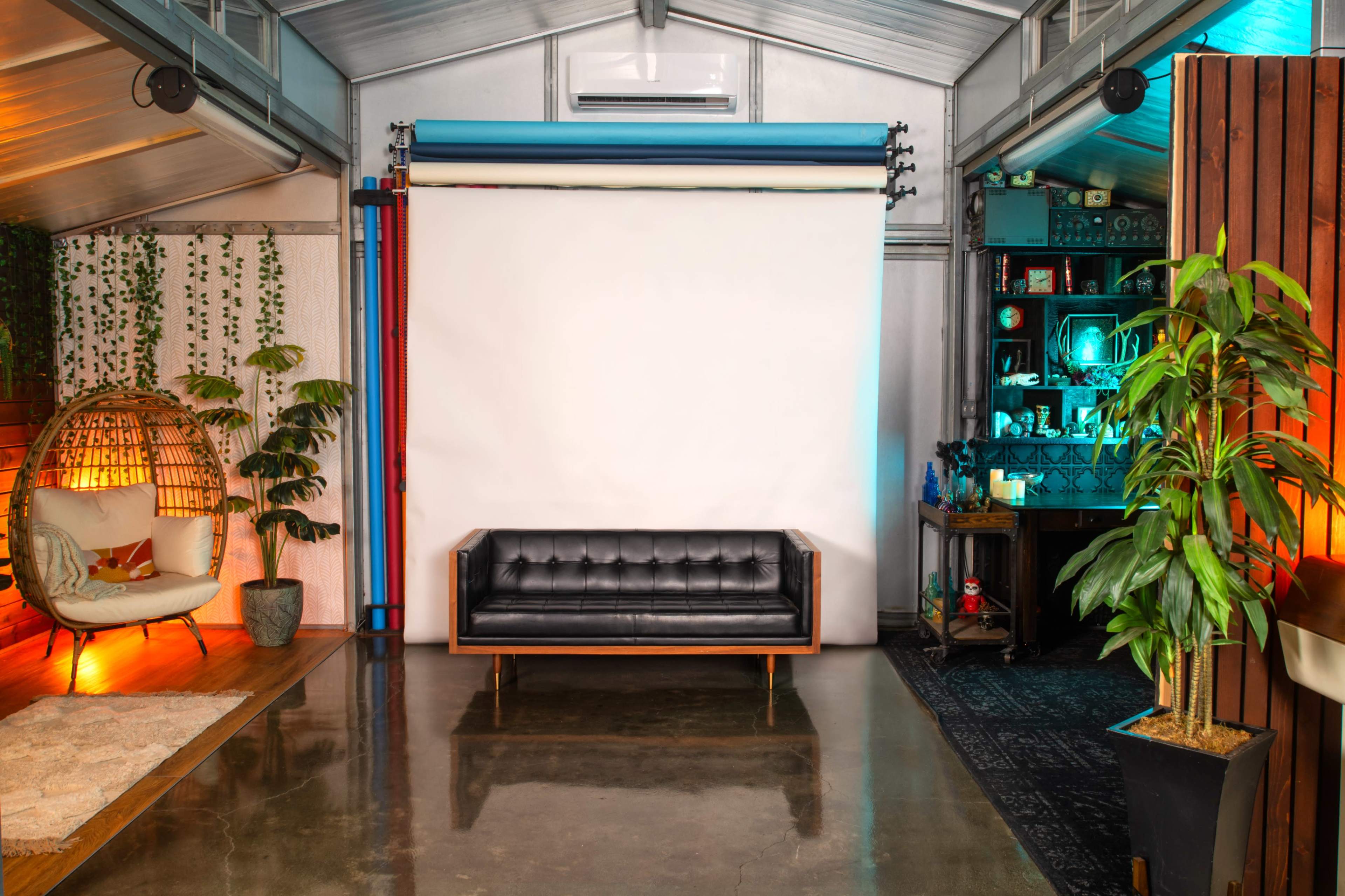 A cozy studio space featuring a black sofa, a light-colored rug, a potted plant, and a backdrop setup against a wall.
