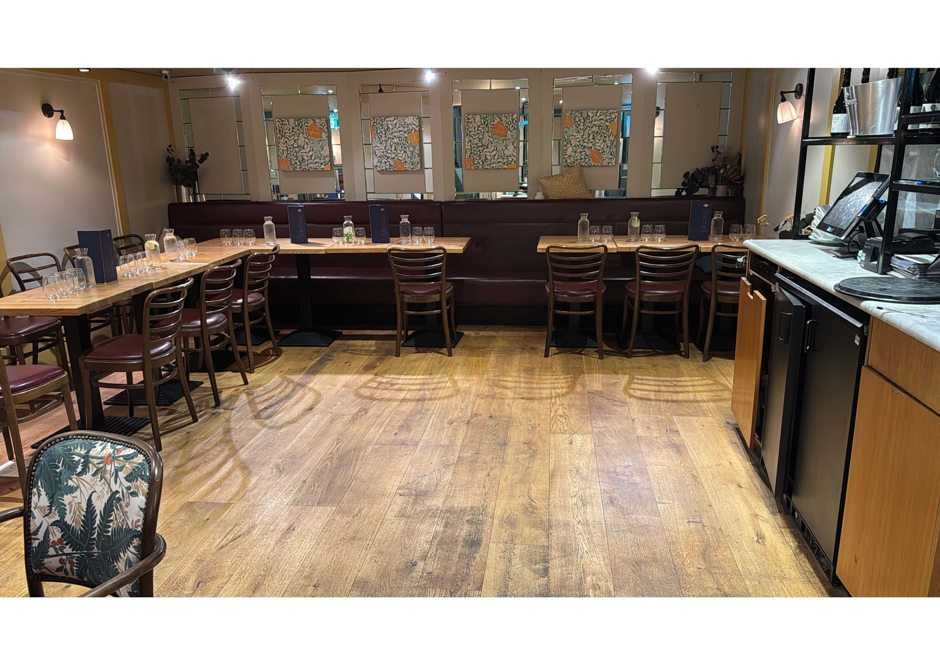 The image shows a restaurant interior with wooden flooring, several tables and chairs, and a bar area on one side.