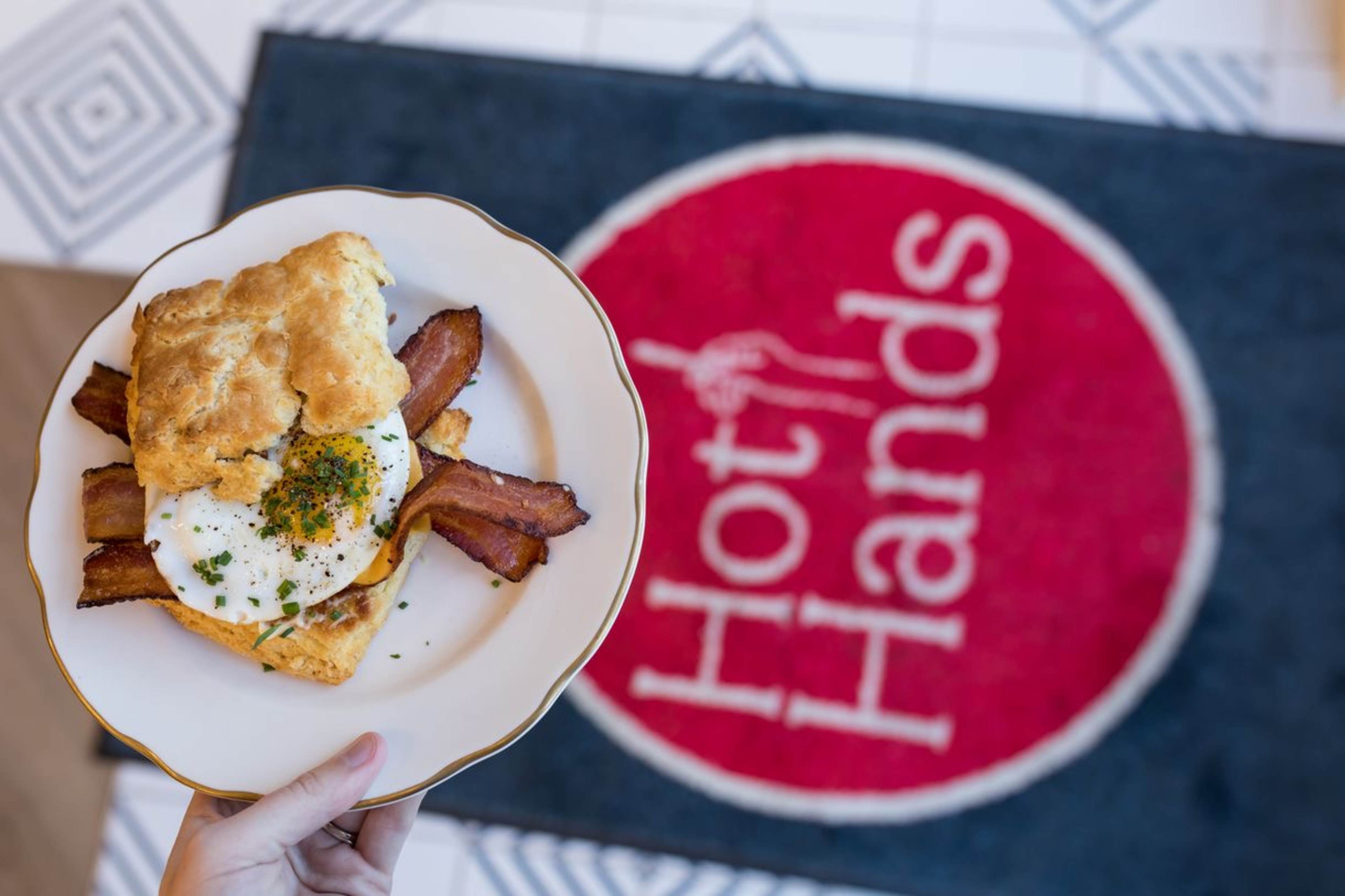 A person holds a plate featuring a biscuit sandwich with bacon and a fried egg in front of a red mat that reads "Hot Hands."