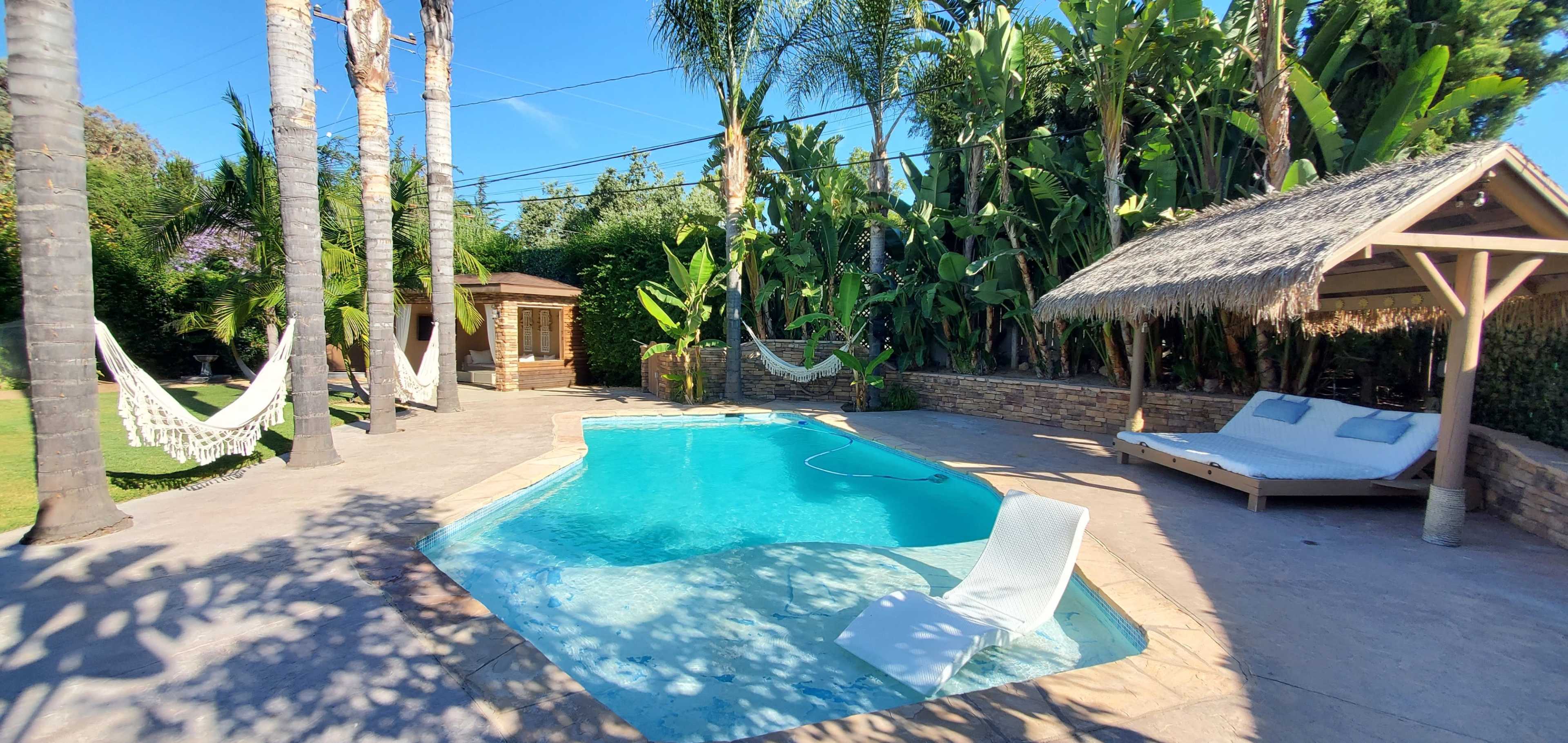 A landscaped backyard with a swimming pool surrounded by palm trees and tropical plants, along with a gazebo and two hammock chairs.