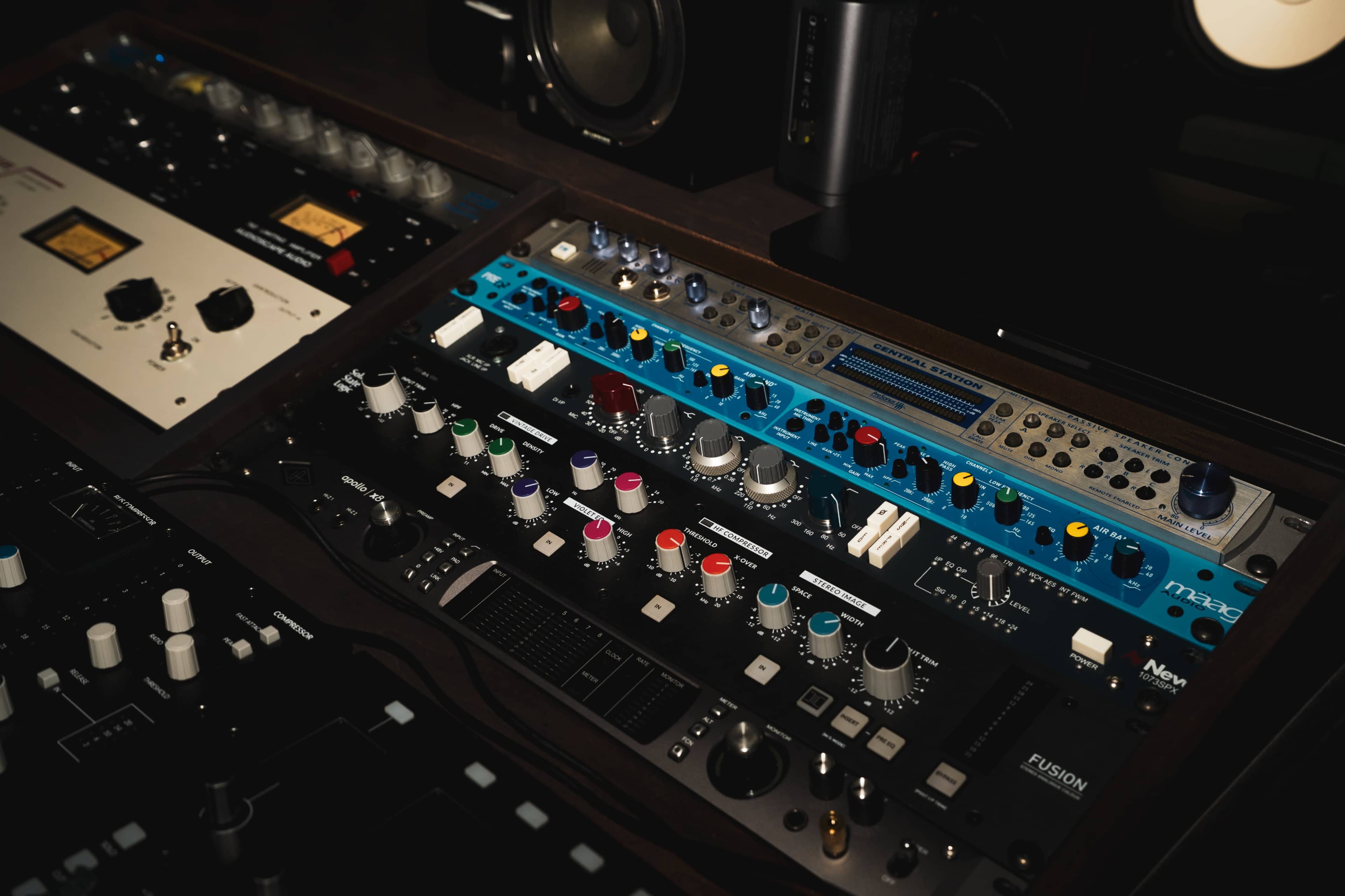 The image shows a close-up view of a sound mixing console featuring various knobs, sliders, and colorful controls, with other audio equipment visible in the background.