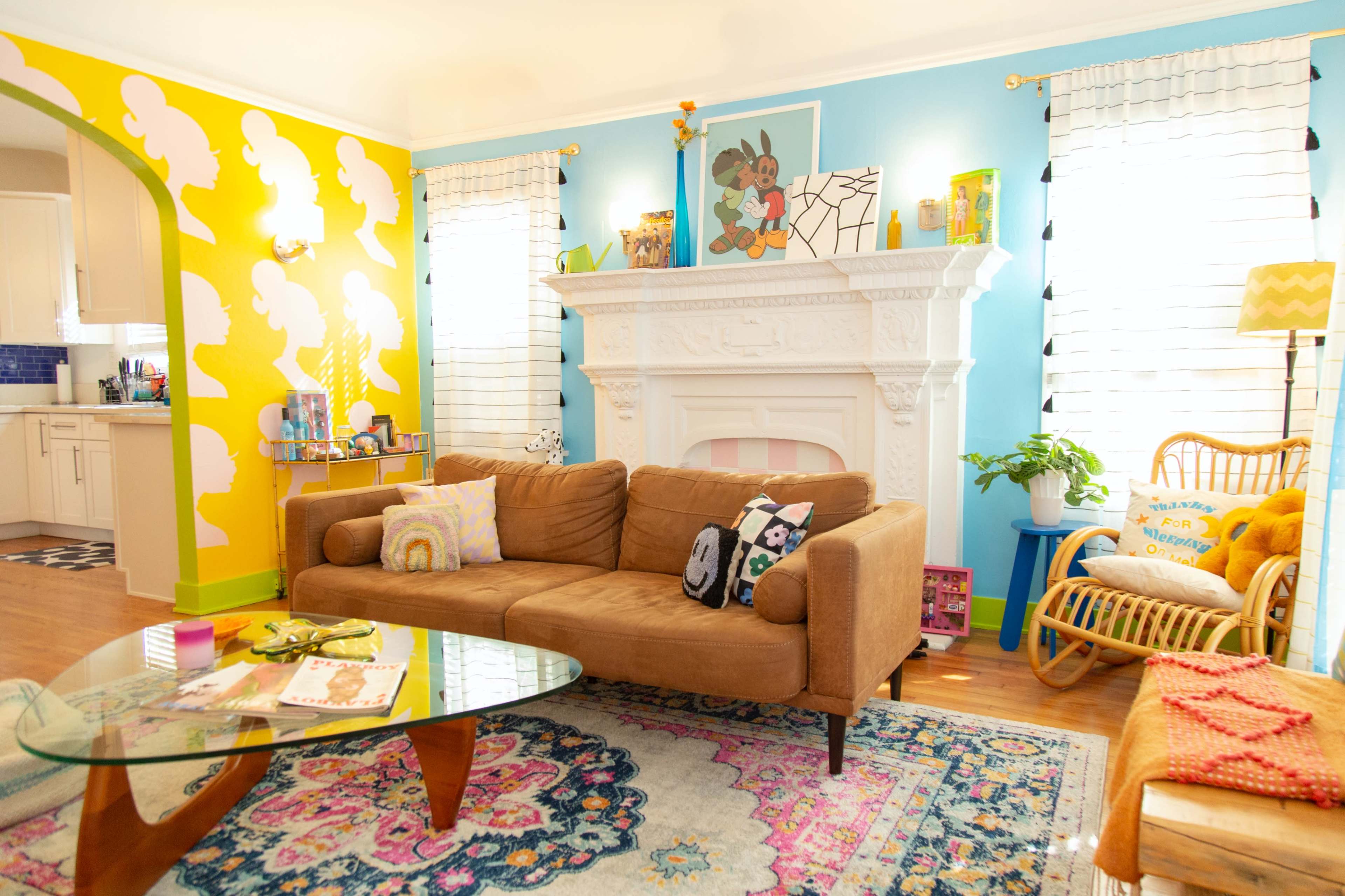 The image shows a brightly decorated living room with a brown sofa, a glass coffee table, colorful wall accents, and various decorative items arranged around a white fireplace.