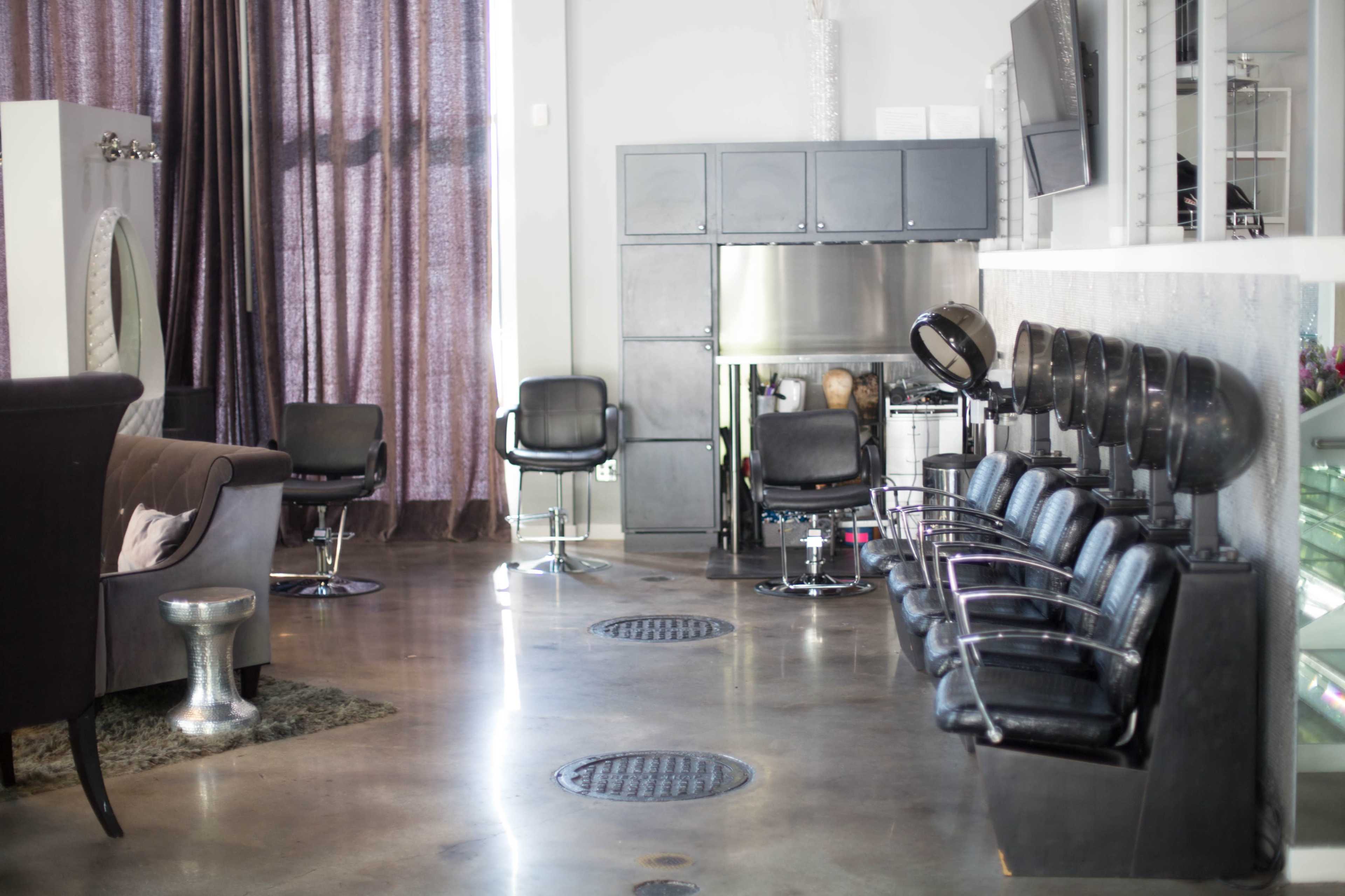 The image shows a modern hair salon with a row of black styling chairs and a seating area featuring a brown couch and large purple curtains.