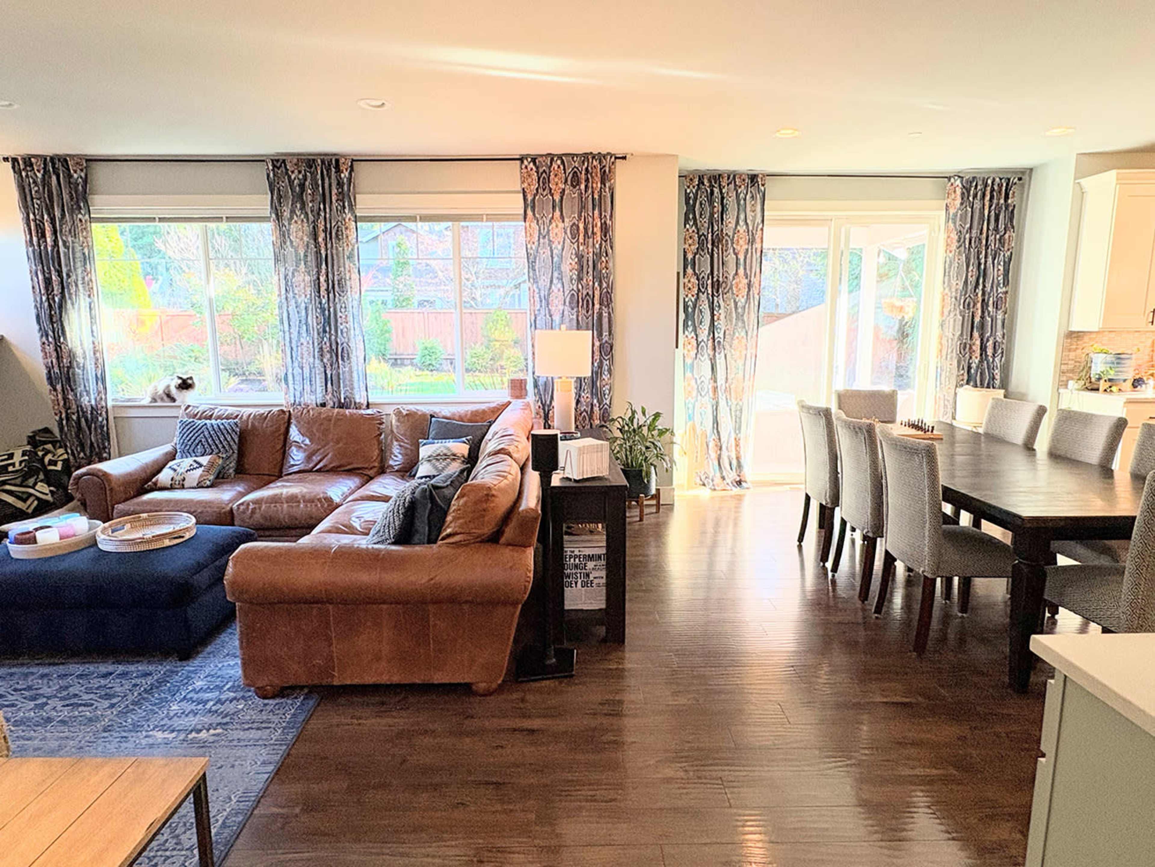 The image shows a spacious living room with a leather sofa and a dining area featuring a long table and several chairs, illuminated by natural light from large windows.
