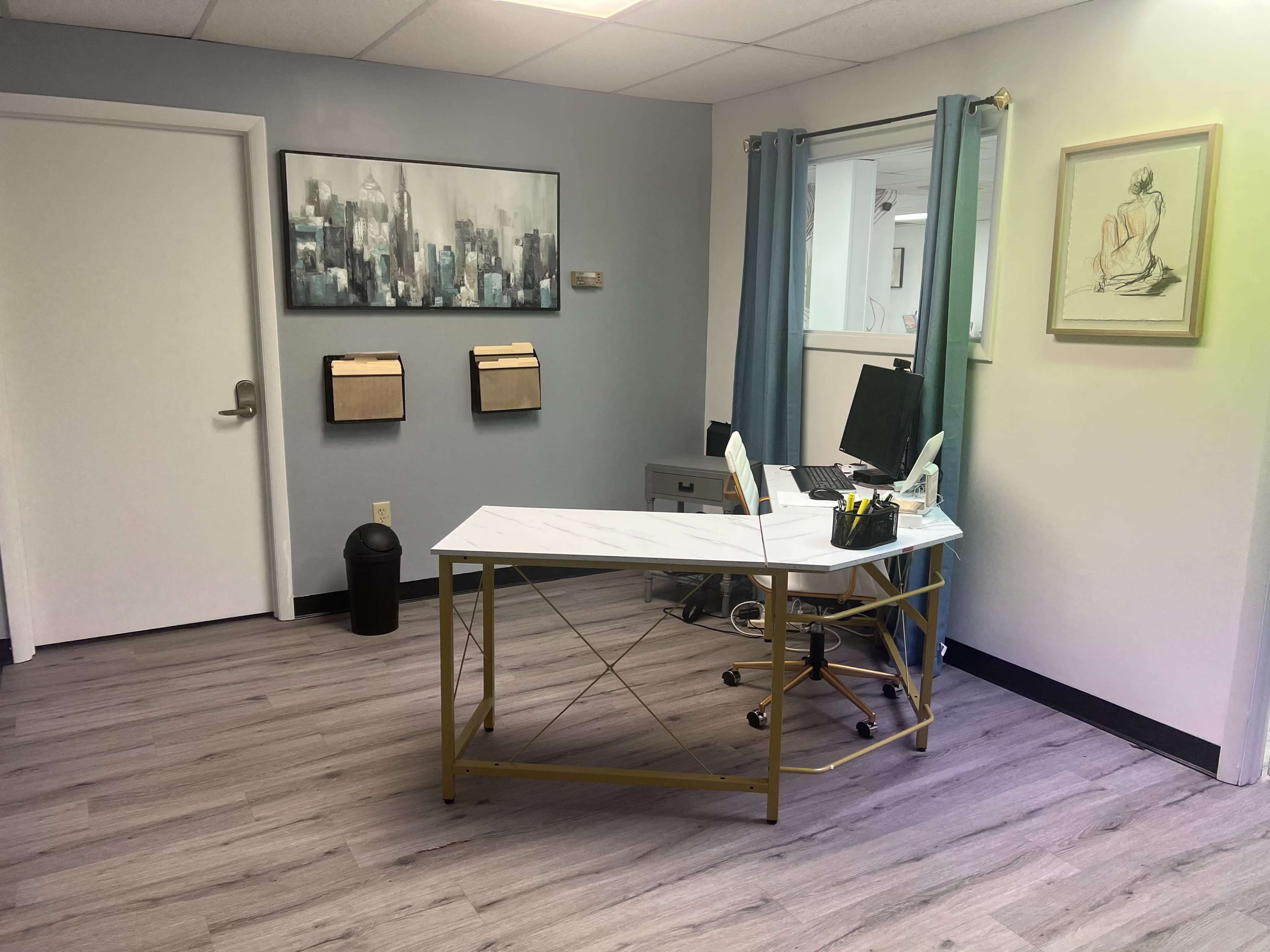 A modern office space featuring a white desk with gold legs, a computer chair, wall-mounted mail organizers, and artwork on the walls.