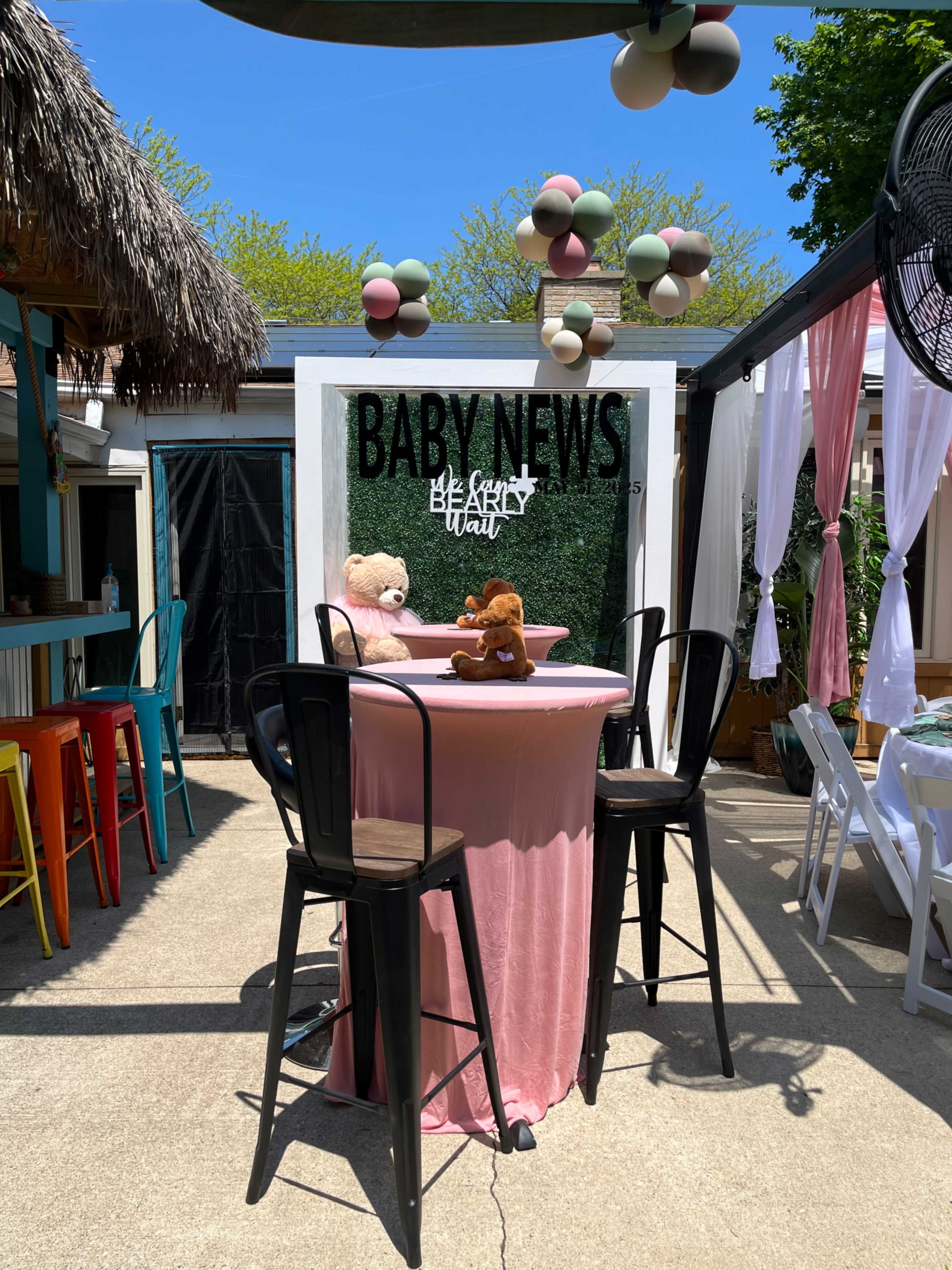 The image shows a decorated outdoor space with a circular table draped in pink, featuring two stuffed animals on top, surrounded by black bar stools and colorful decor overhead.