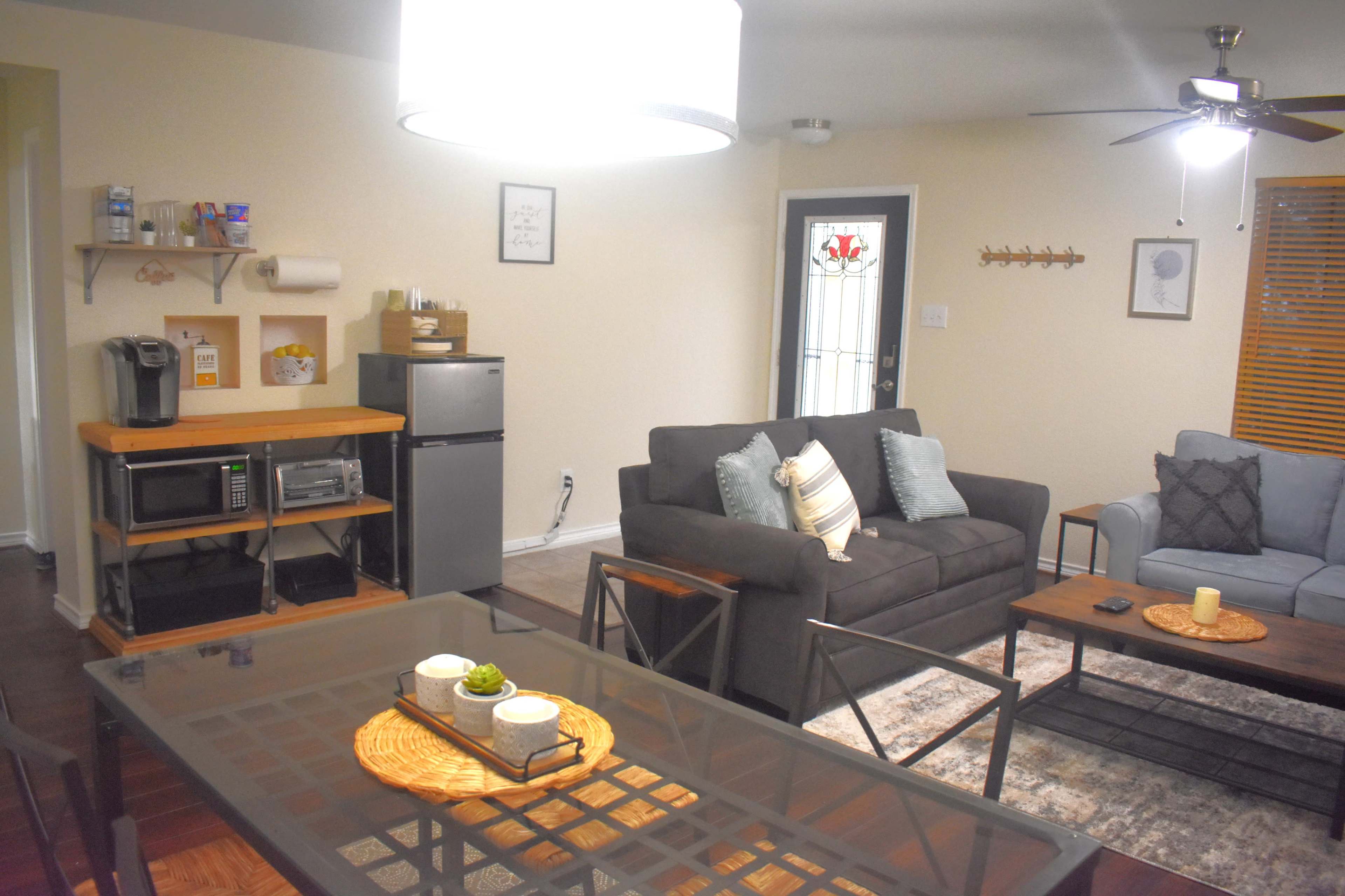 The image shows a cozy living space with a dining area, a gray sofa, a blue armchair, a small kitchen setup with appliances, and a door leading outside.