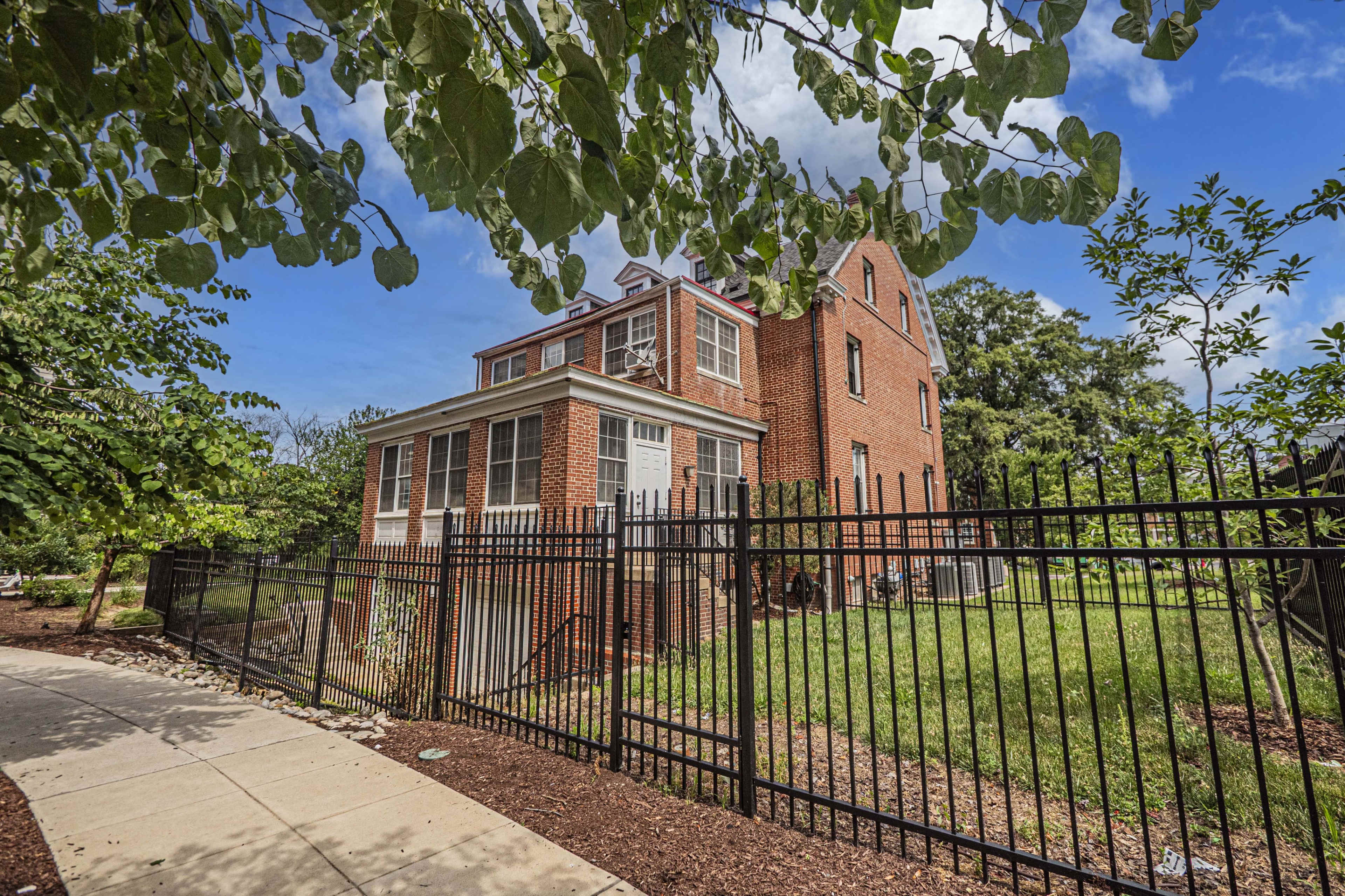 A three-story red brick house is enclosed by a black metal fence and surrounded by greenery, with a sidewalk leading up to it.