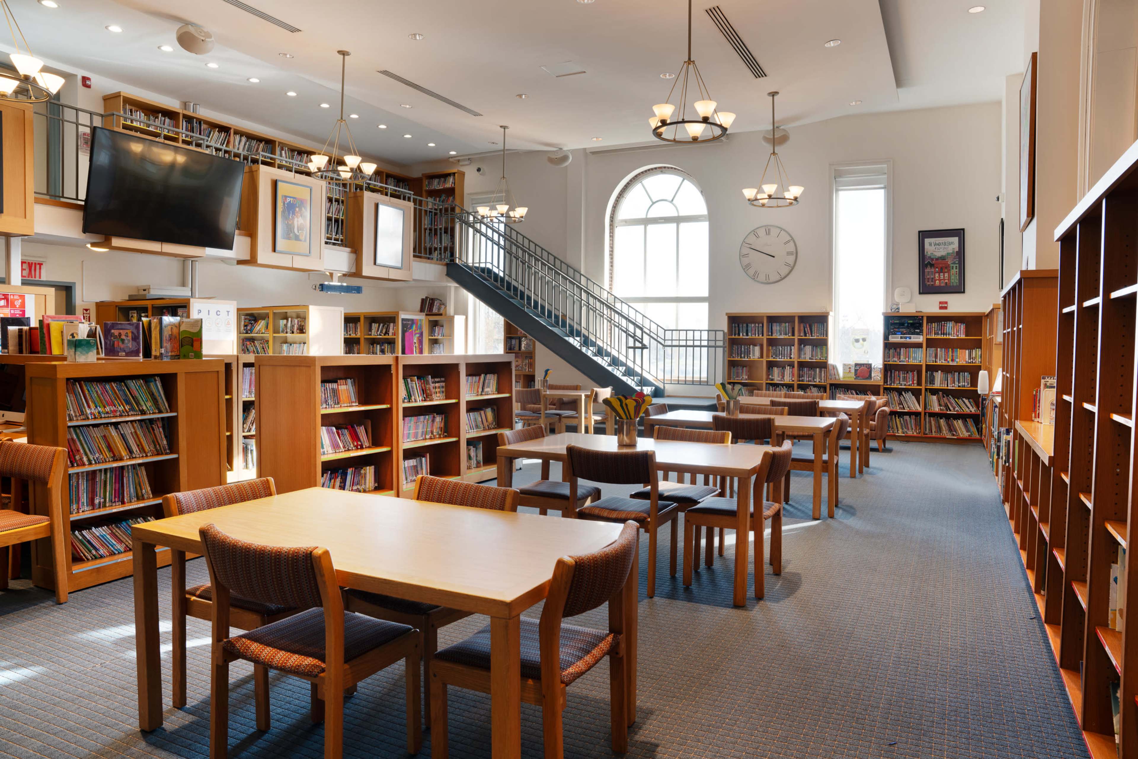 A spacious library with wooden shelves filled with books, several tables and chairs, and a staircase leading to an upper level.