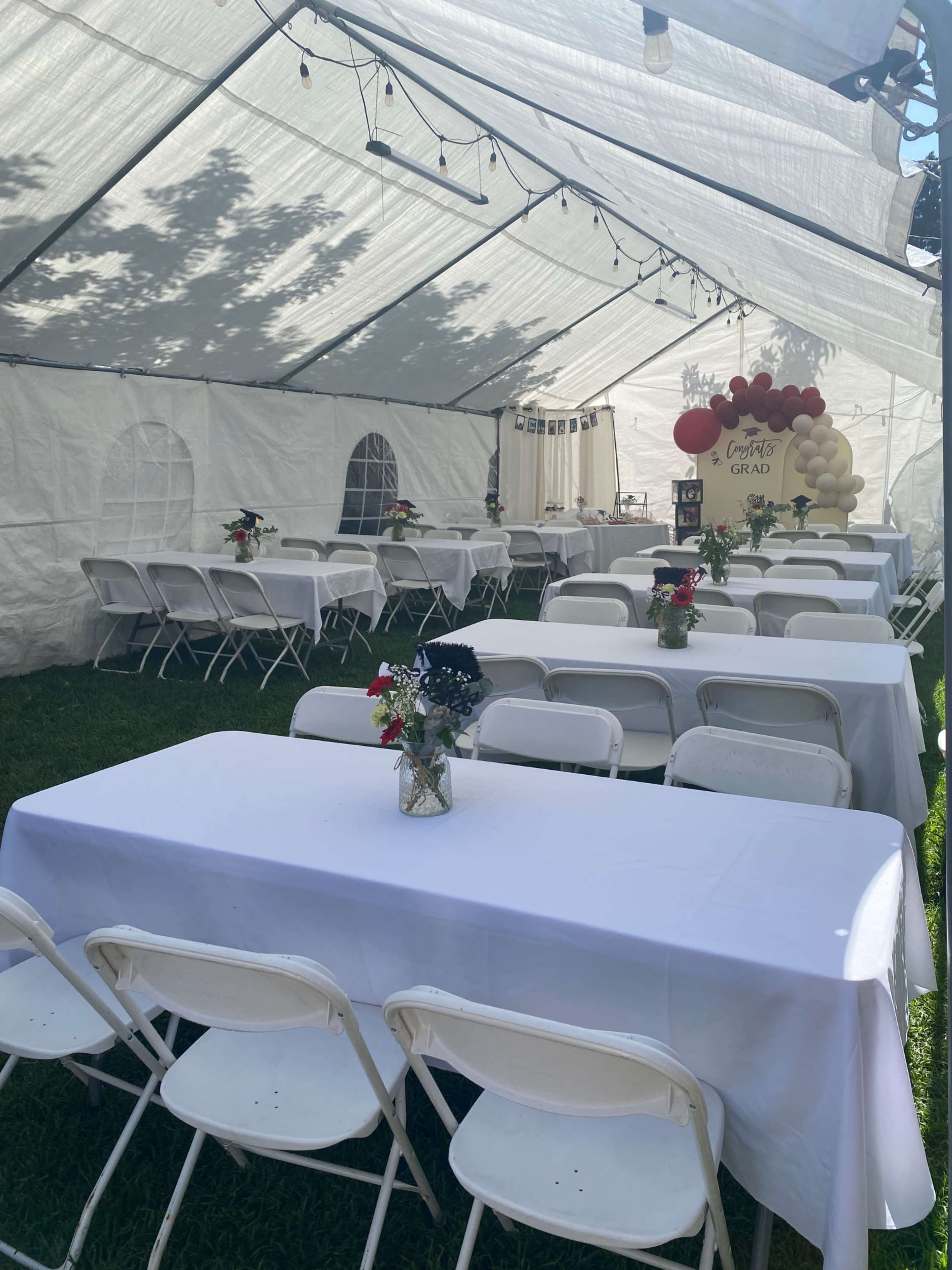 A large white tent is set up outdoors, containing rows of tables covered with white tablecloths and decorated with small floral centerpieces, alongside balloon decorations for a graduation event.