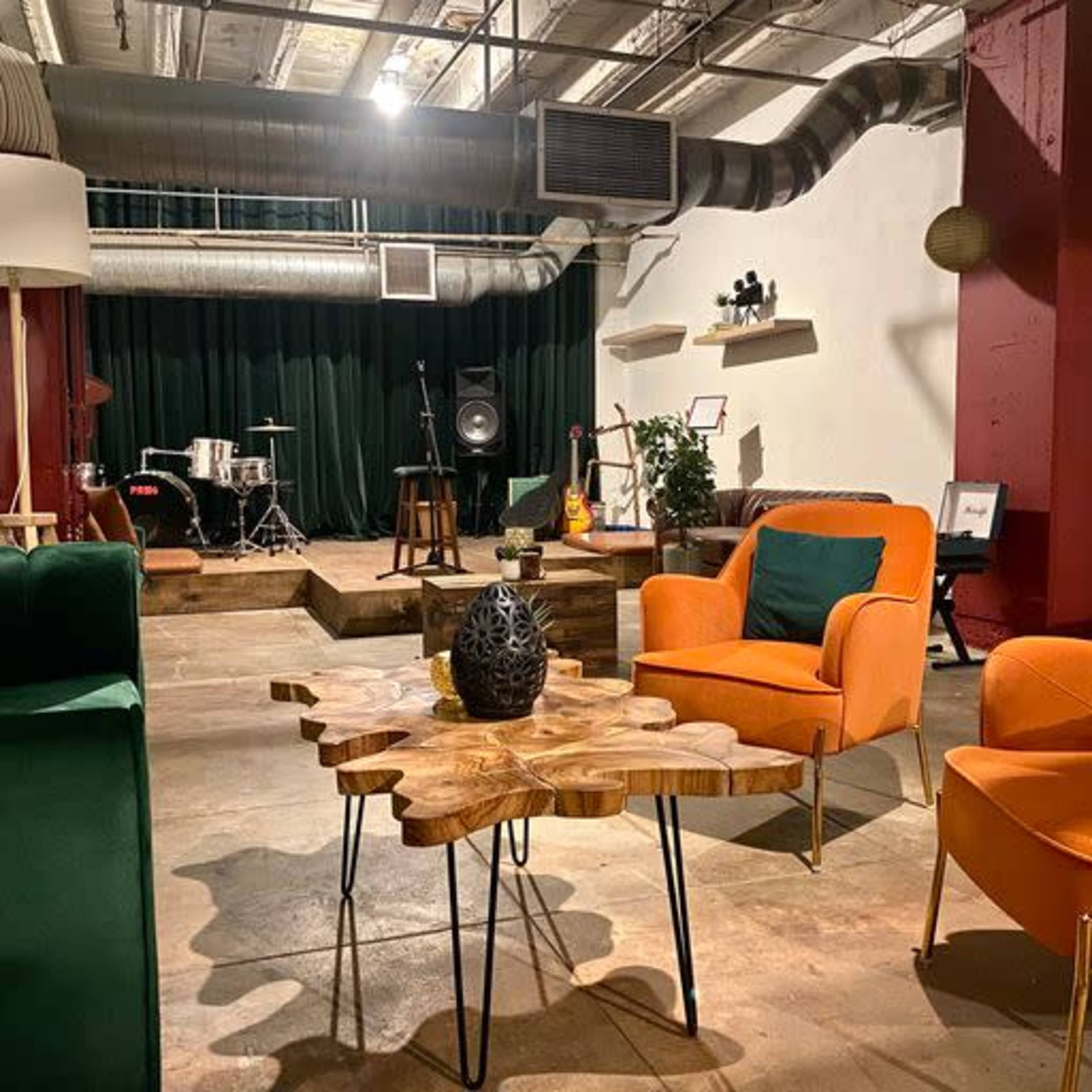 A modern lounge area with orange chairs, a wooden coffee table, and various musical instruments set up in a spacious room with exposed ventilation and industrial decor.