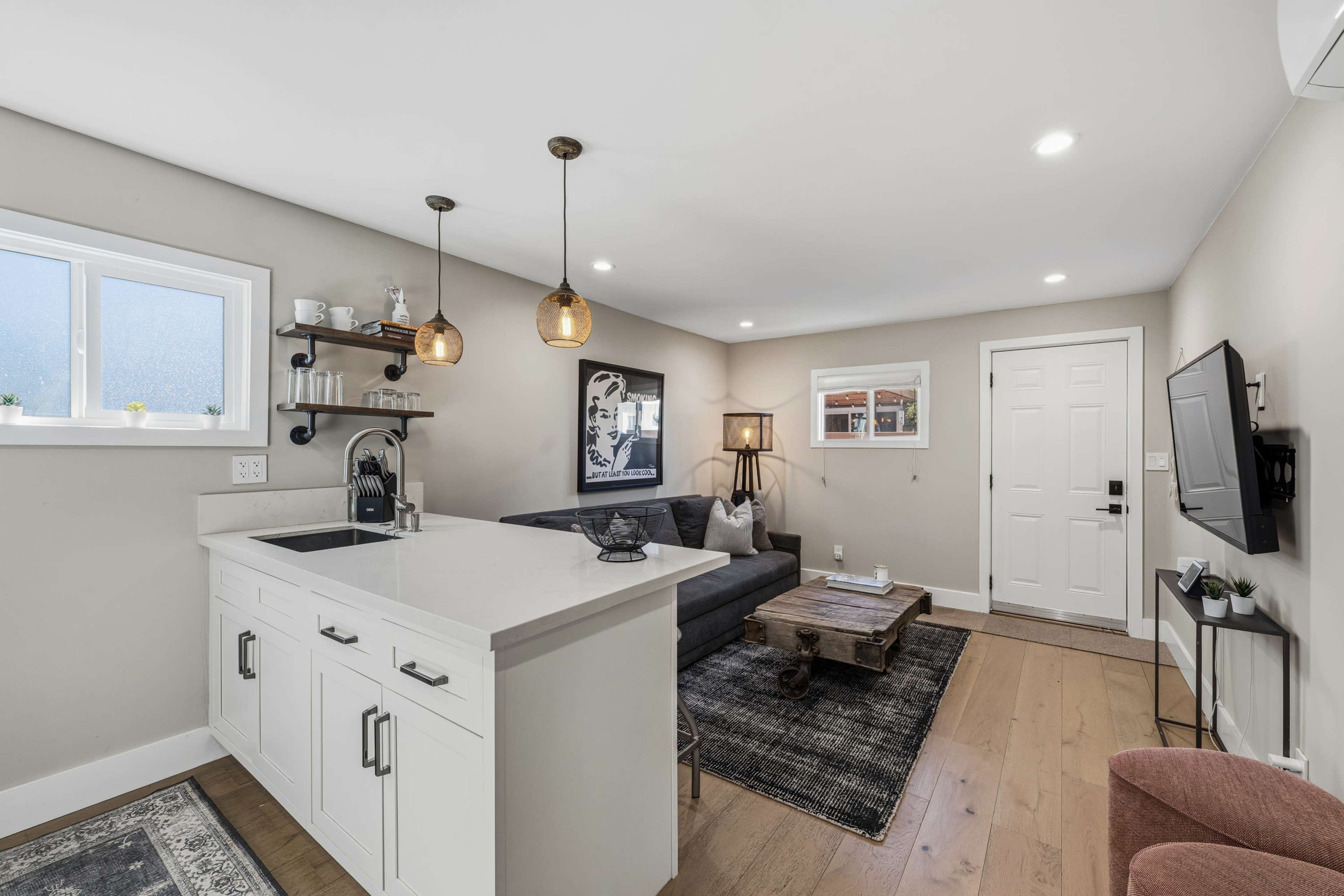 The image shows a modern living space with a kitchen area featuring a countertop, a gray sectional sofa, a wooden coffee table, and a front door with a small window.