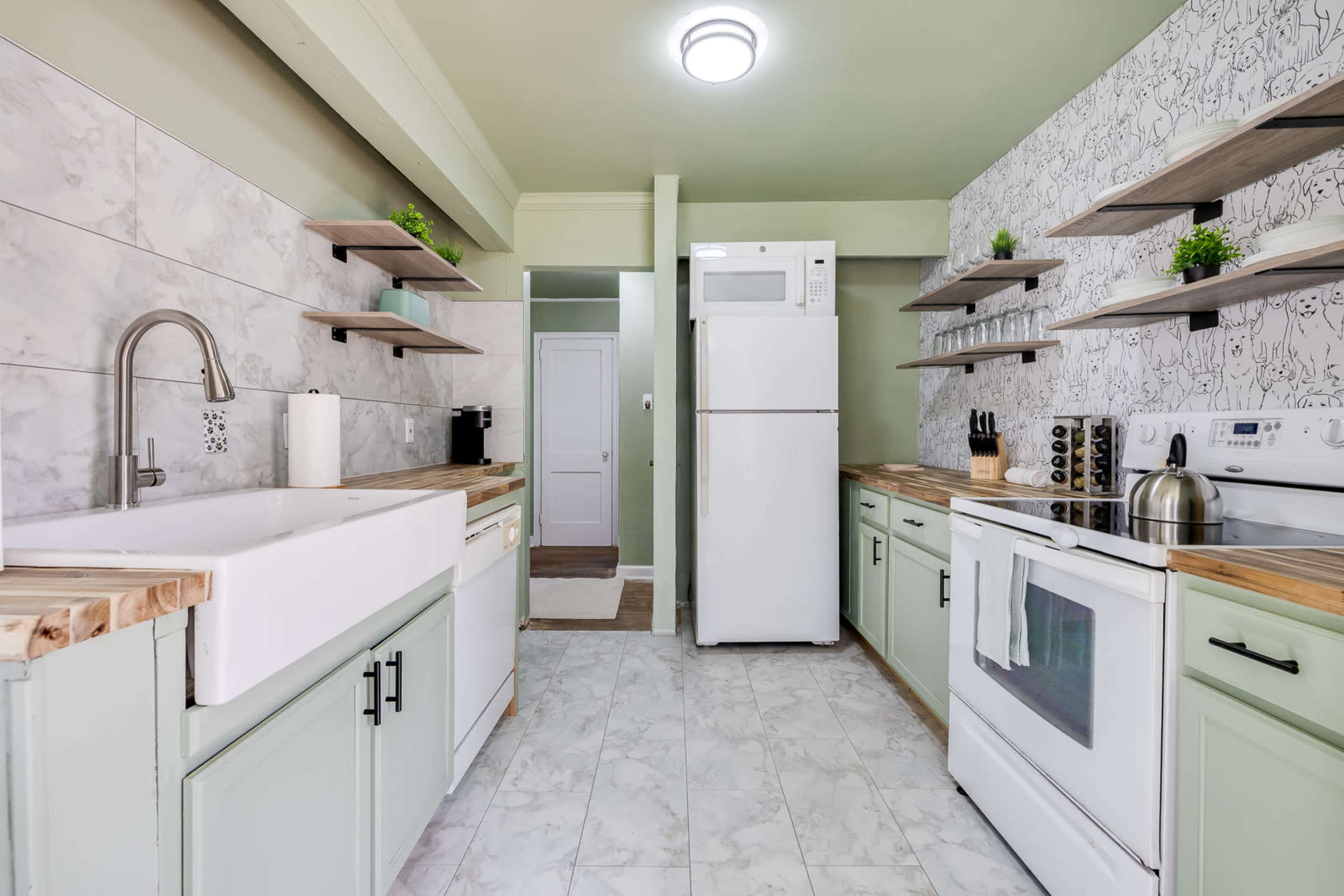 The image depicts a modern kitchen featuring light green cabinetry, a white farmhouse sink, and stainless steel appliances set against a backdrop of patterned wallpaper and wood shelving.