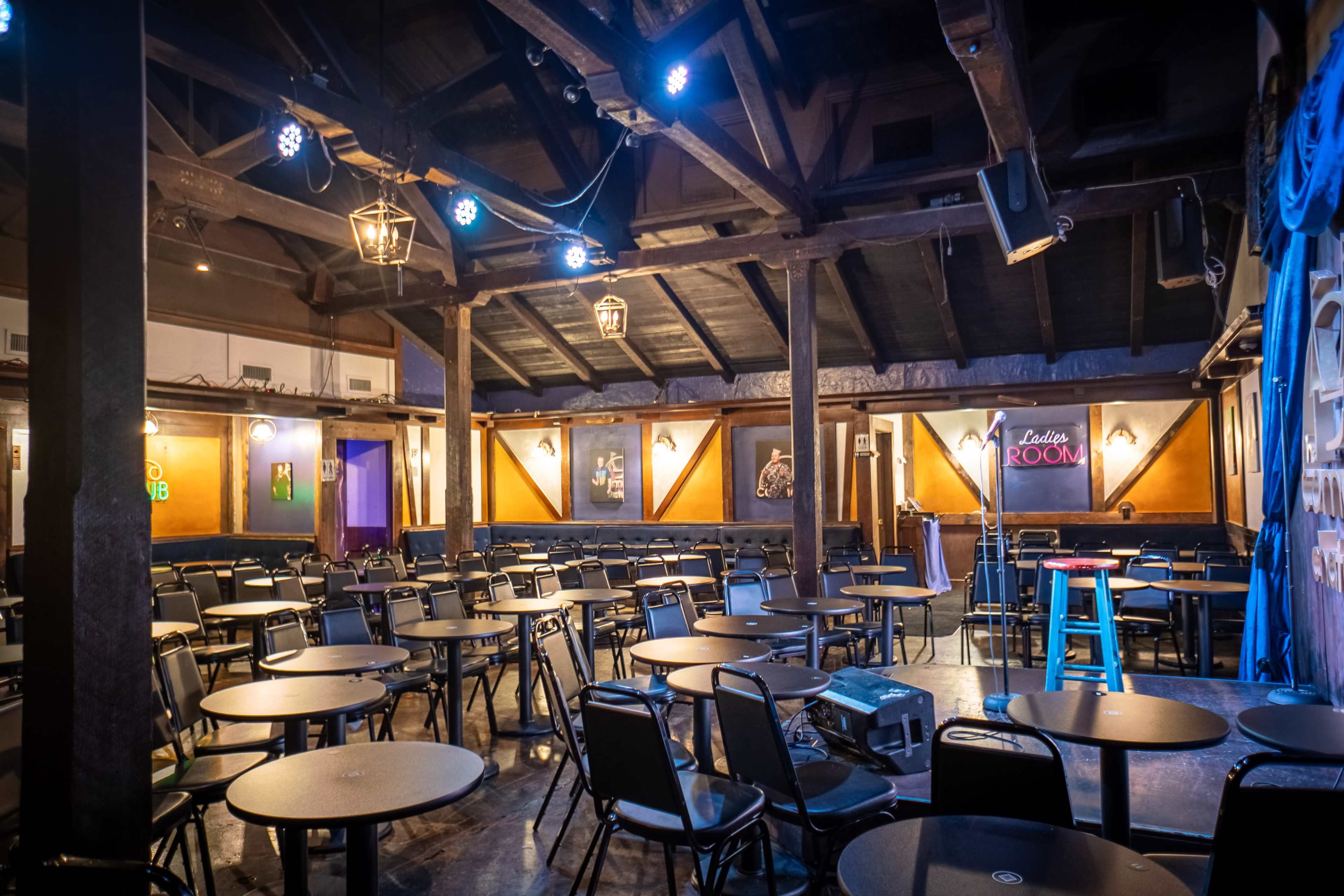 An interior view of a performance space with rows of tables and chairs set up under wooden beams and dim lighting.