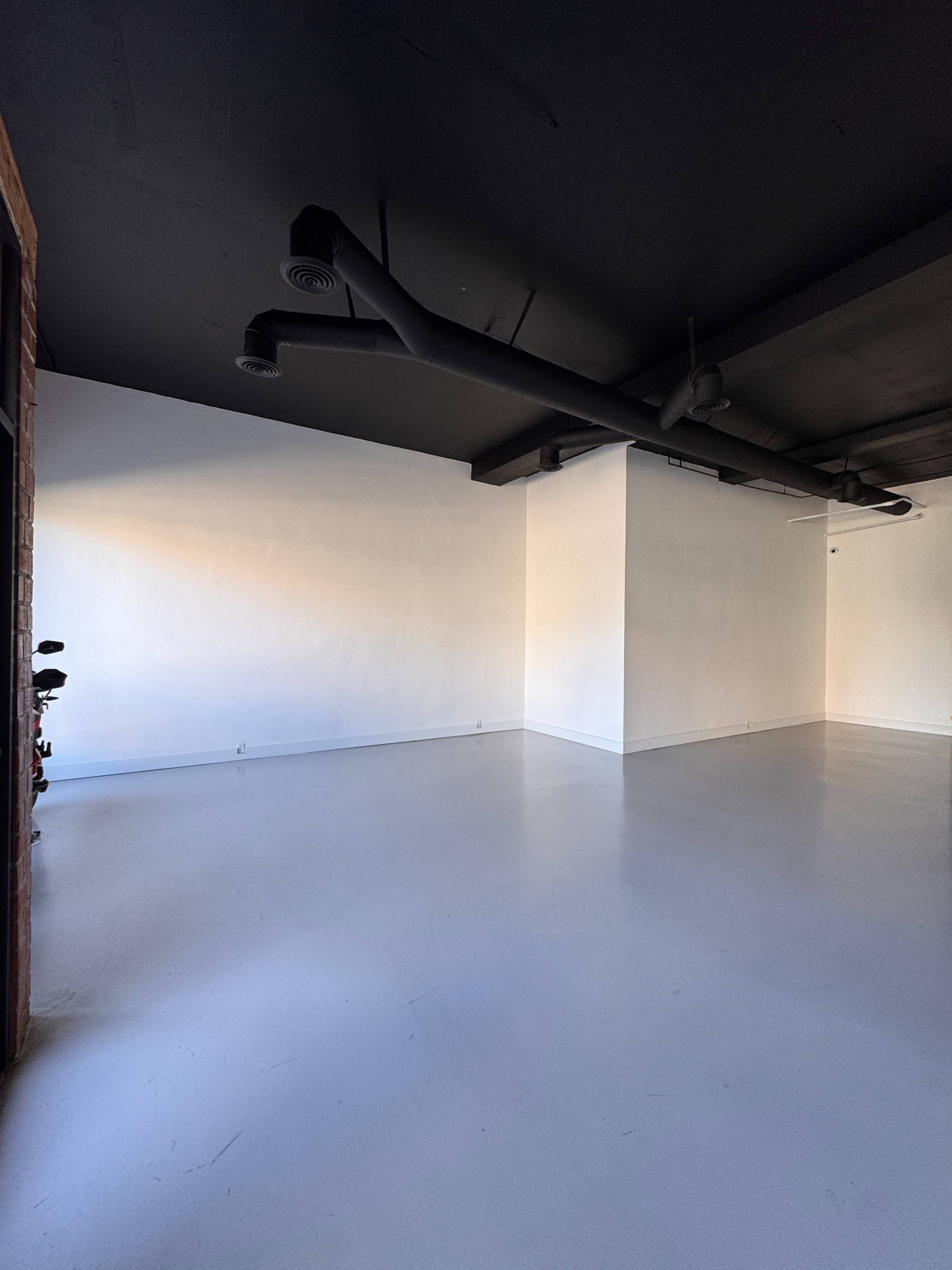The image shows a spacious, empty room with white walls and a gray ceiling, featuring exposed pipes along the ceiling and a smooth, light-colored floor.