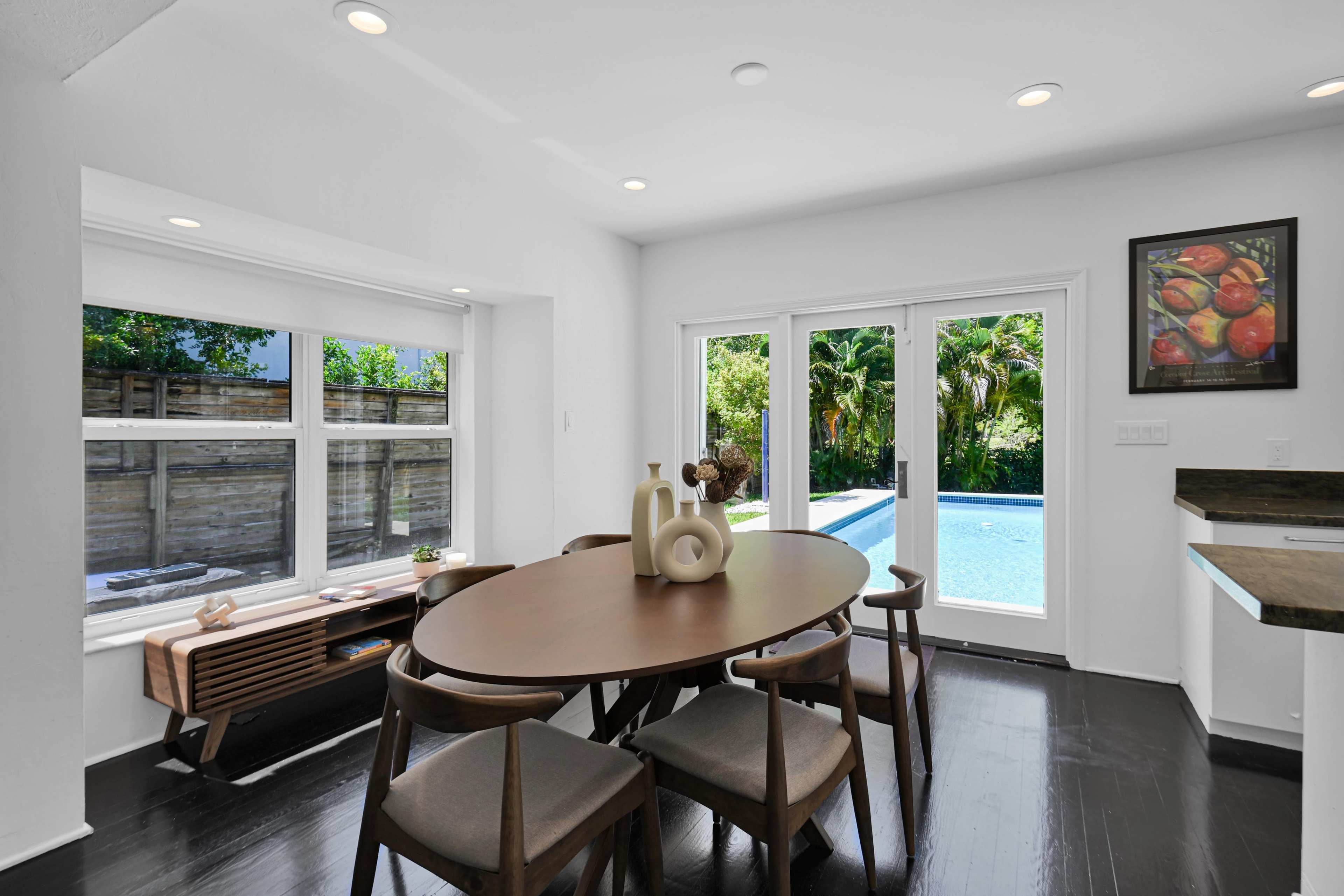 A dining area with a round table and wooden chairs overlooks a patio and pool area through glass doors.