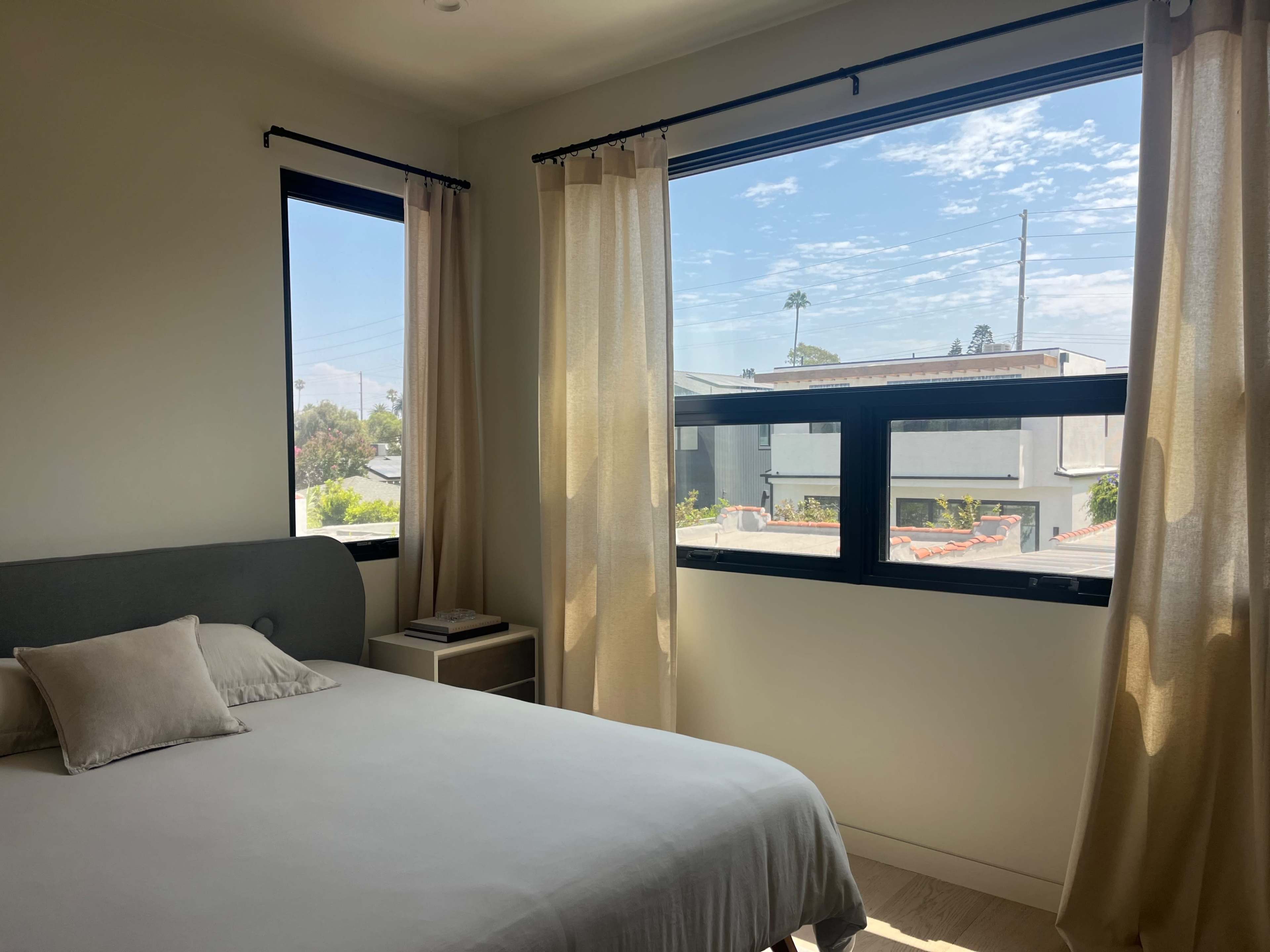 A bright bedroom features a bed with a light-colored duvet and two windows dressed with sheer curtains, allowing natural light to illuminate the space.