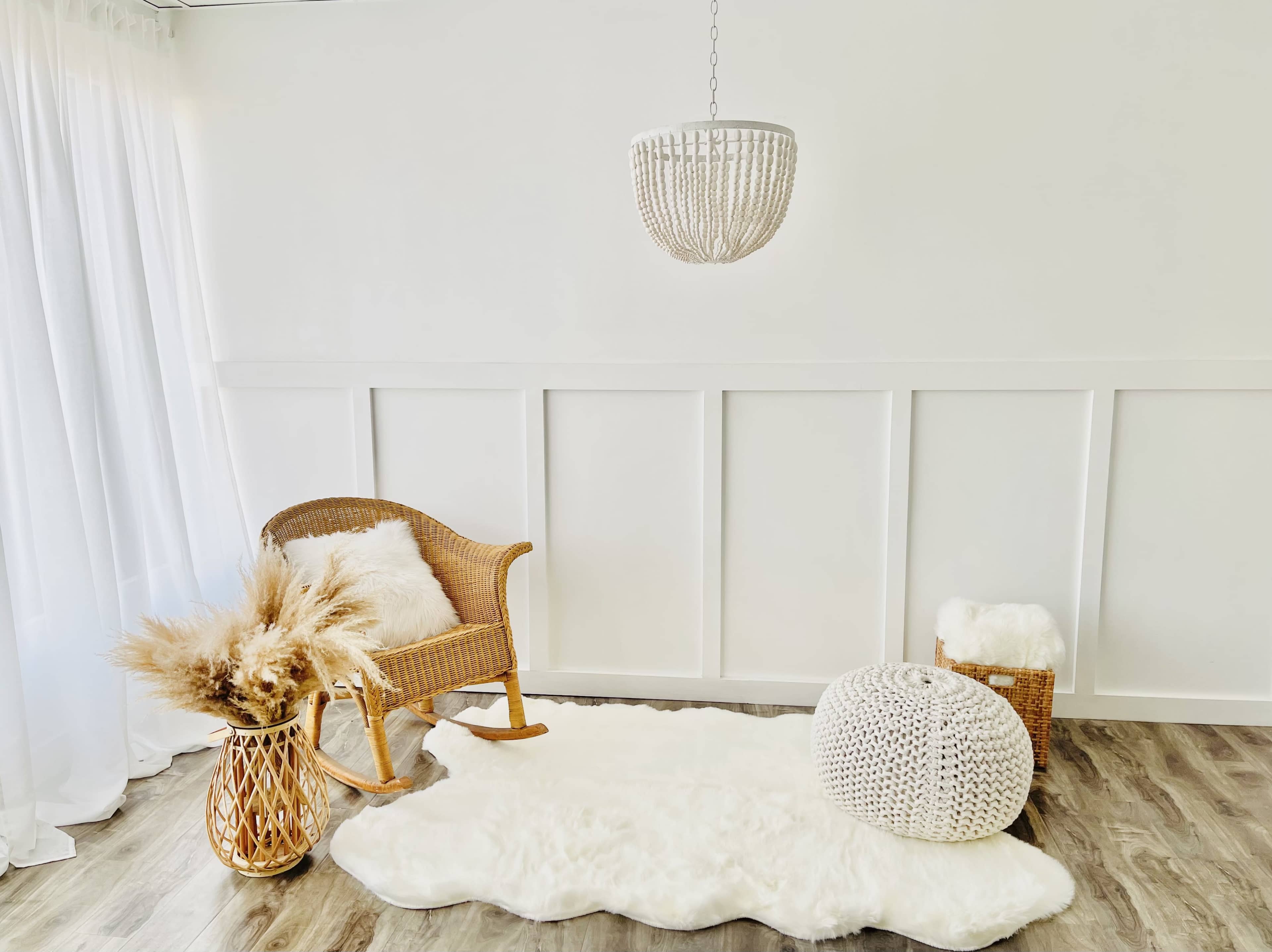 A cozy interior corner with a wicker rocking chair, a hanging light fixture, a plush white rug, a decorative pouf, and a basket holding pampas grass.