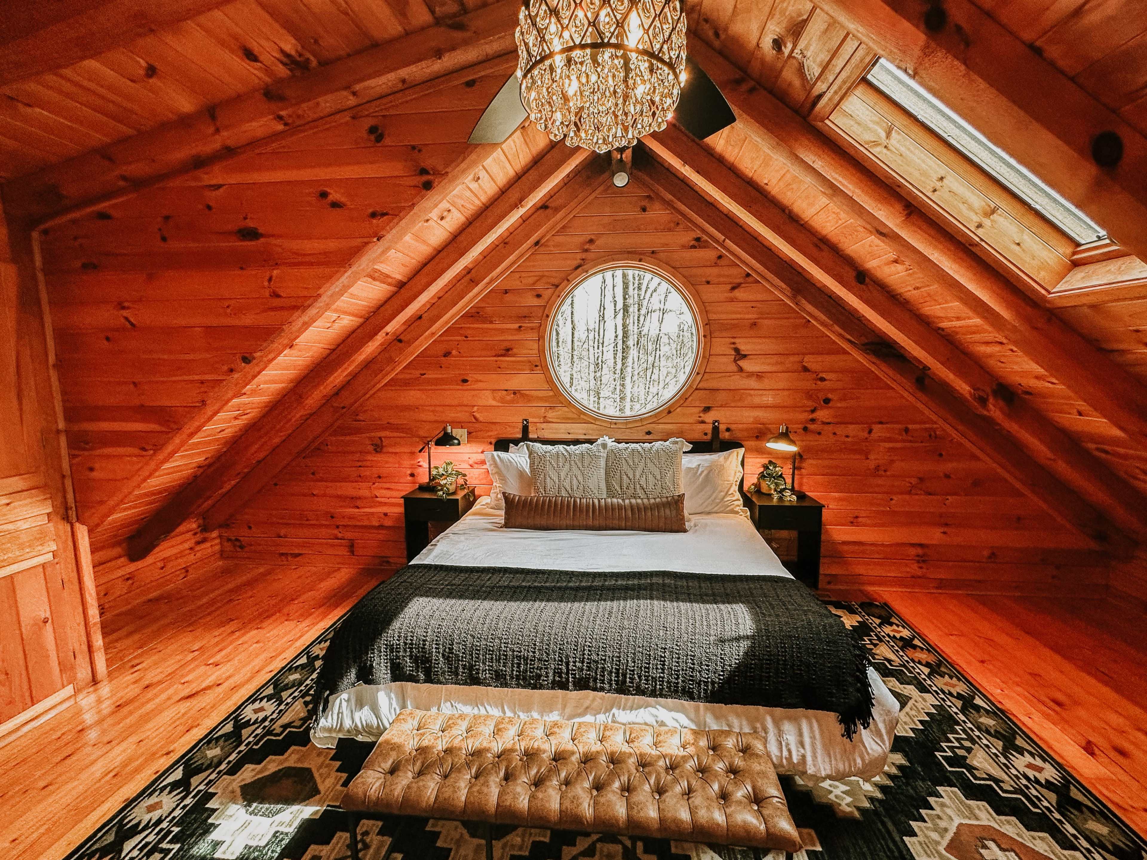 A cozy attic bedroom features a king-sized bed with a dark blanket, flanked by nightstands, and has a round window and a skylight above.
