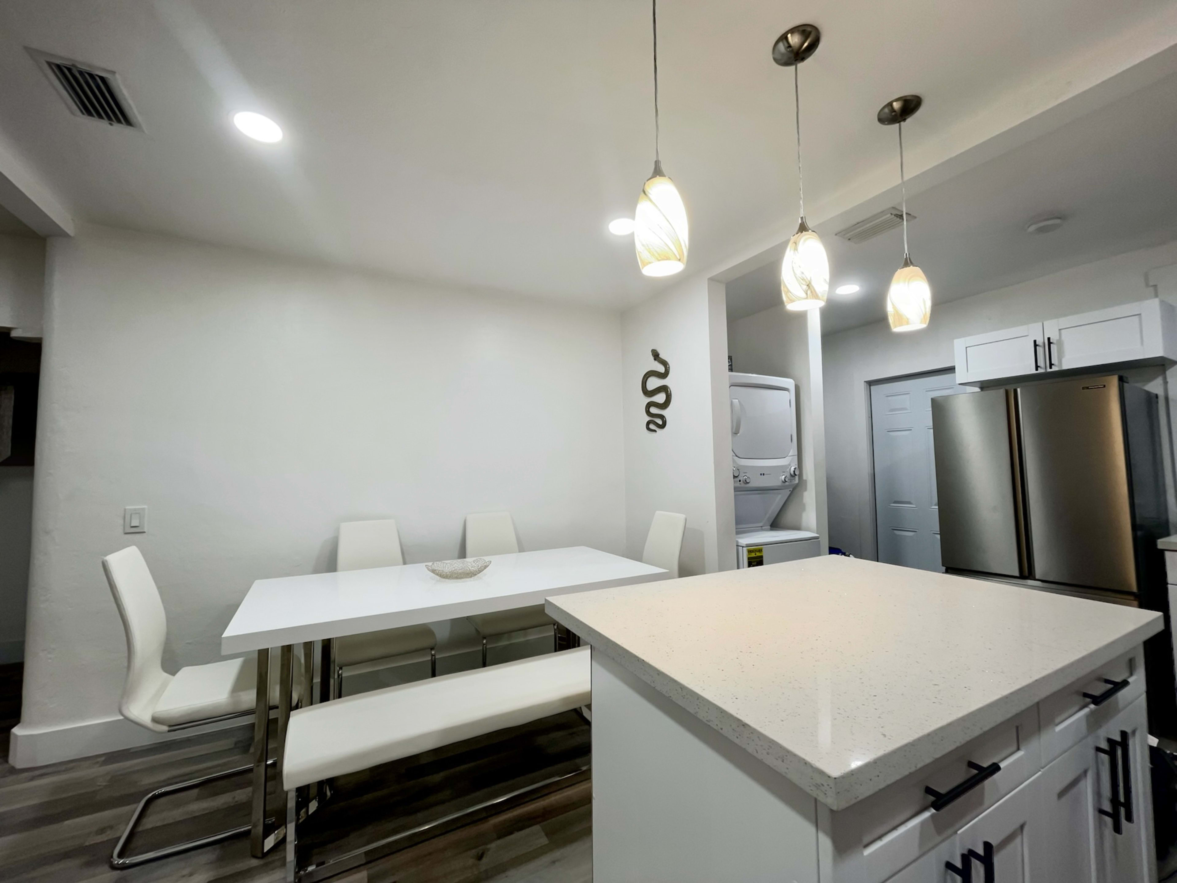 The image shows a modern kitchen and dining area with a white table, six chairs, stainless steel appliances, and pendant lighting.