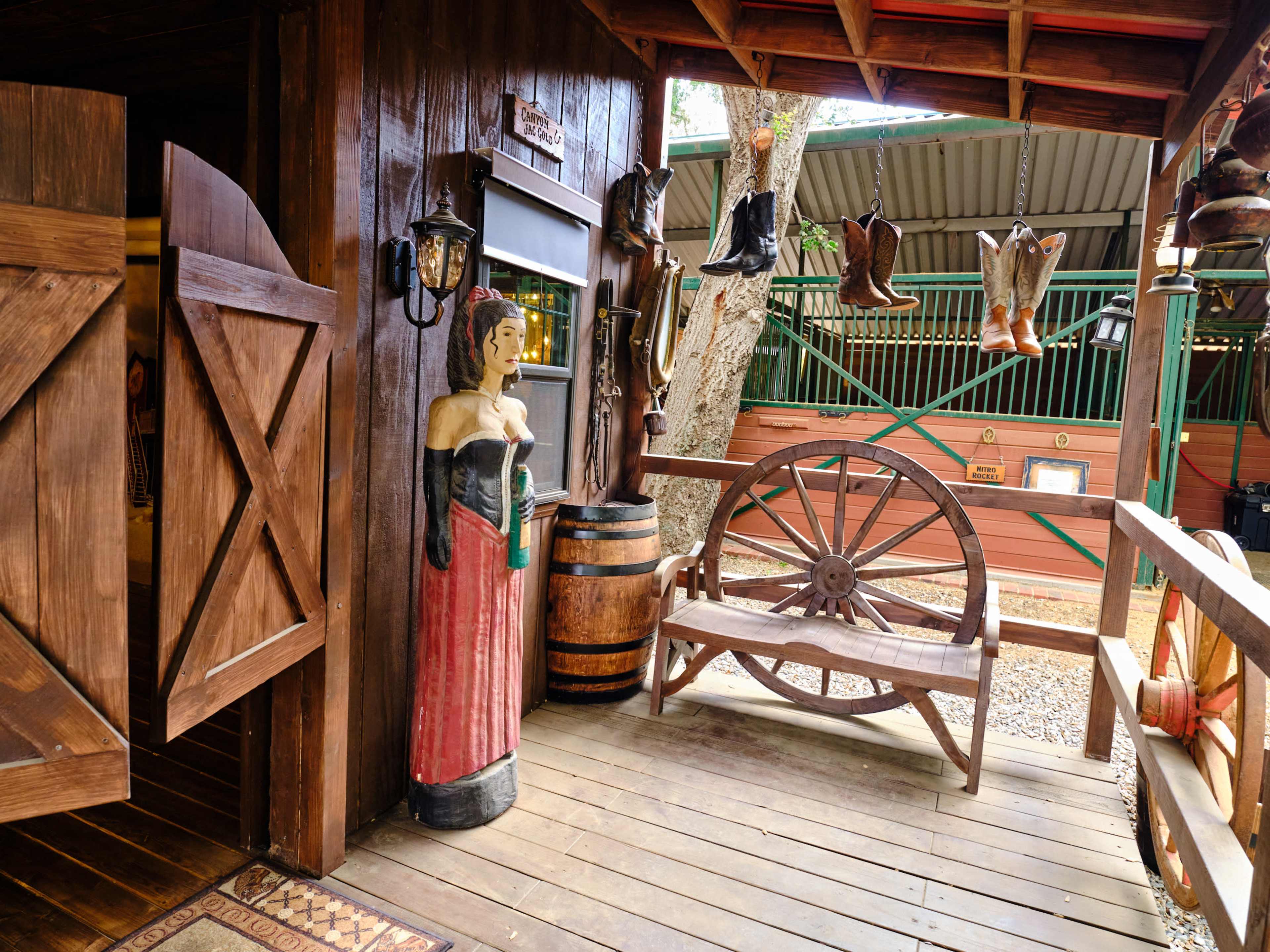 The scene features a rustic wooden entrance with a carved figure, a barrel, a wagon wheel bench, and several pairs of boots hanging on the wall.