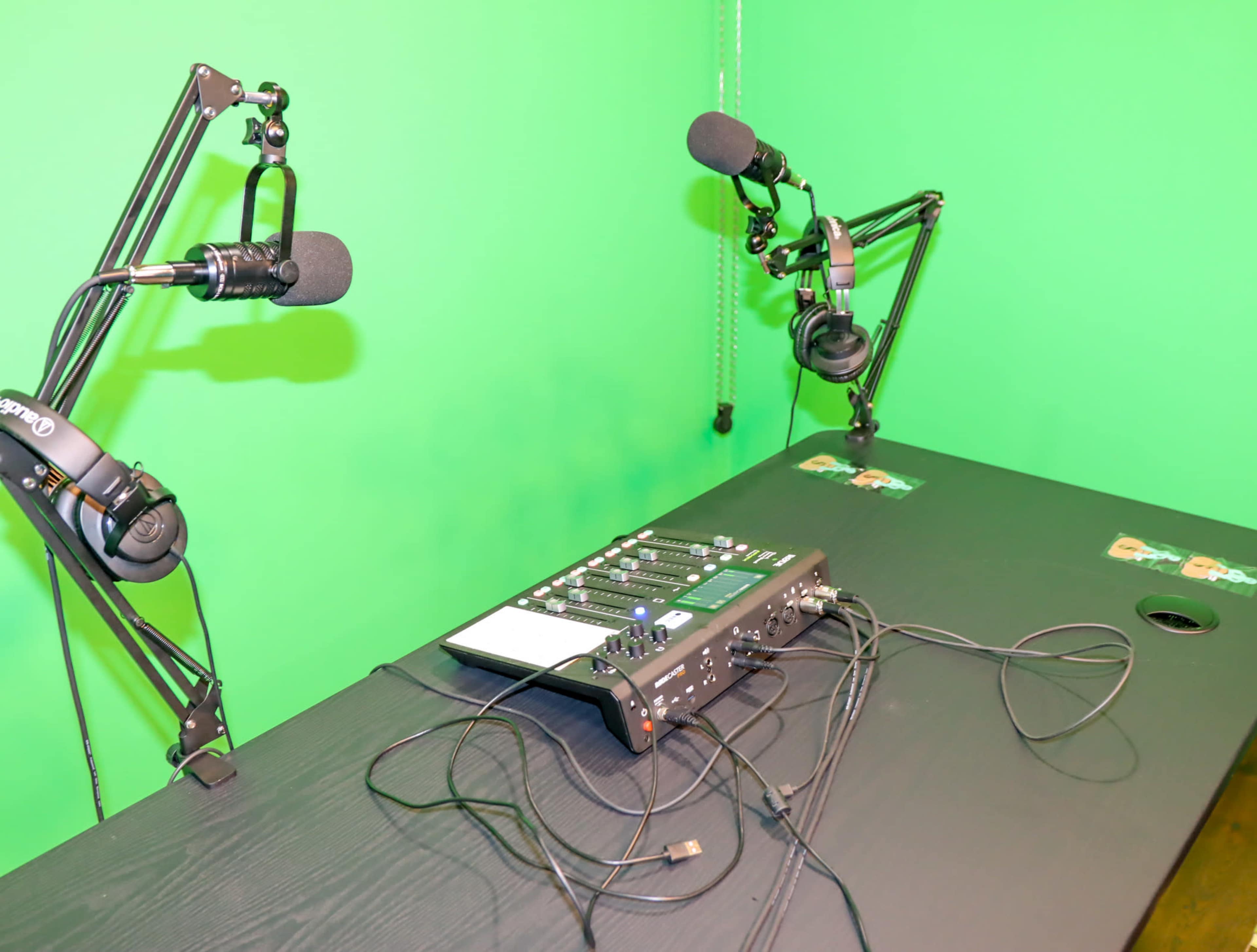 A black table with microphones mounted on adjustable arms and an audio mixer is placed in front of a green screen.