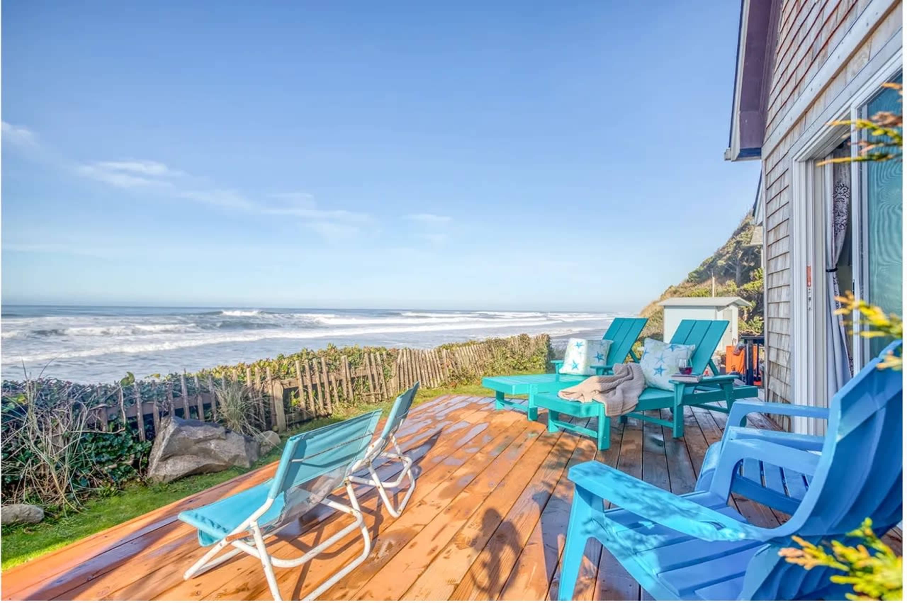 A coastal deck features colorful chairs overlooking a beach with rolling waves and a clear sky.