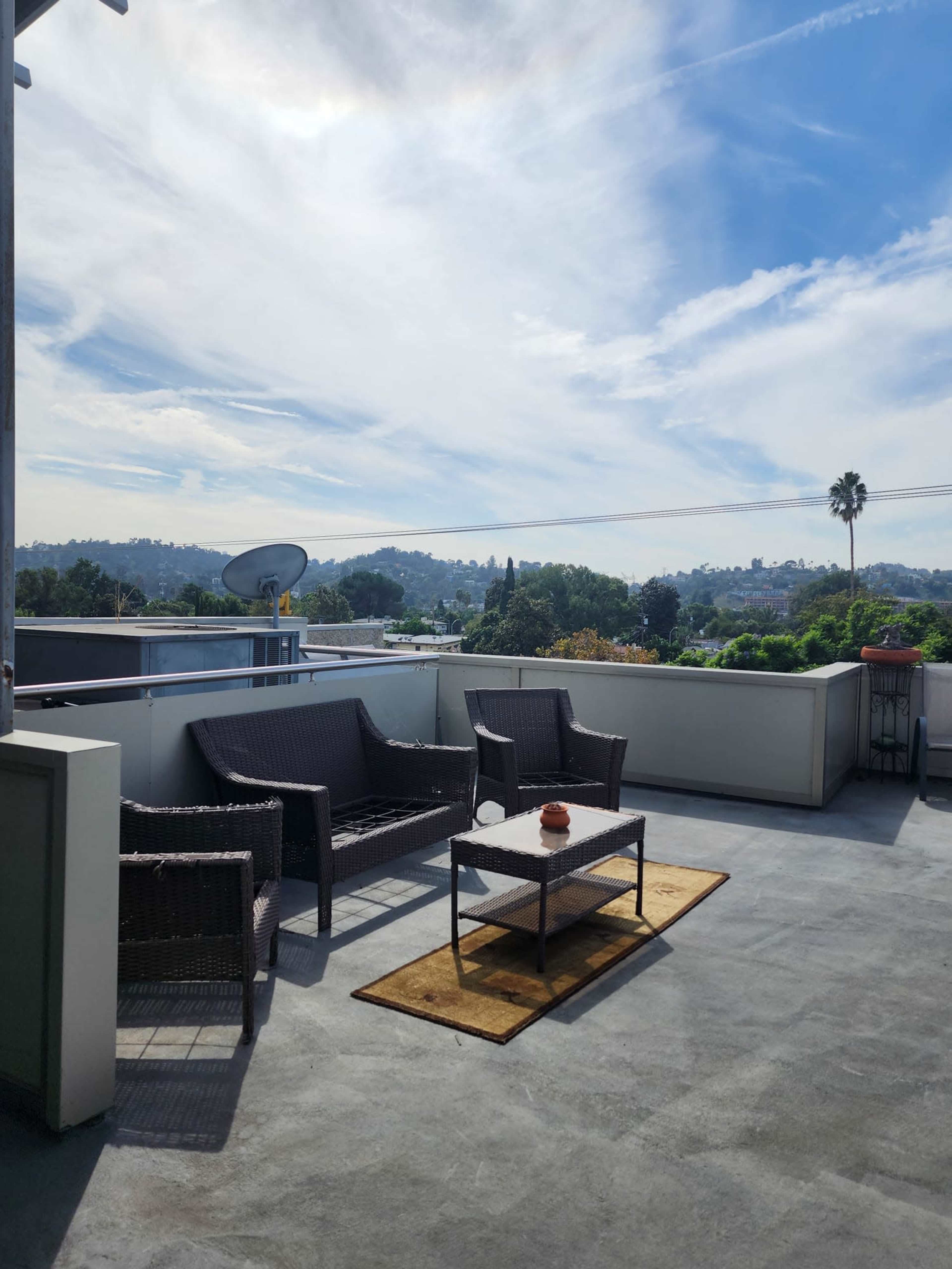 A modern outdoor terrace features wicker furniture, a table with a small centerpiece, and views of hills and trees under a partly cloudy sky.