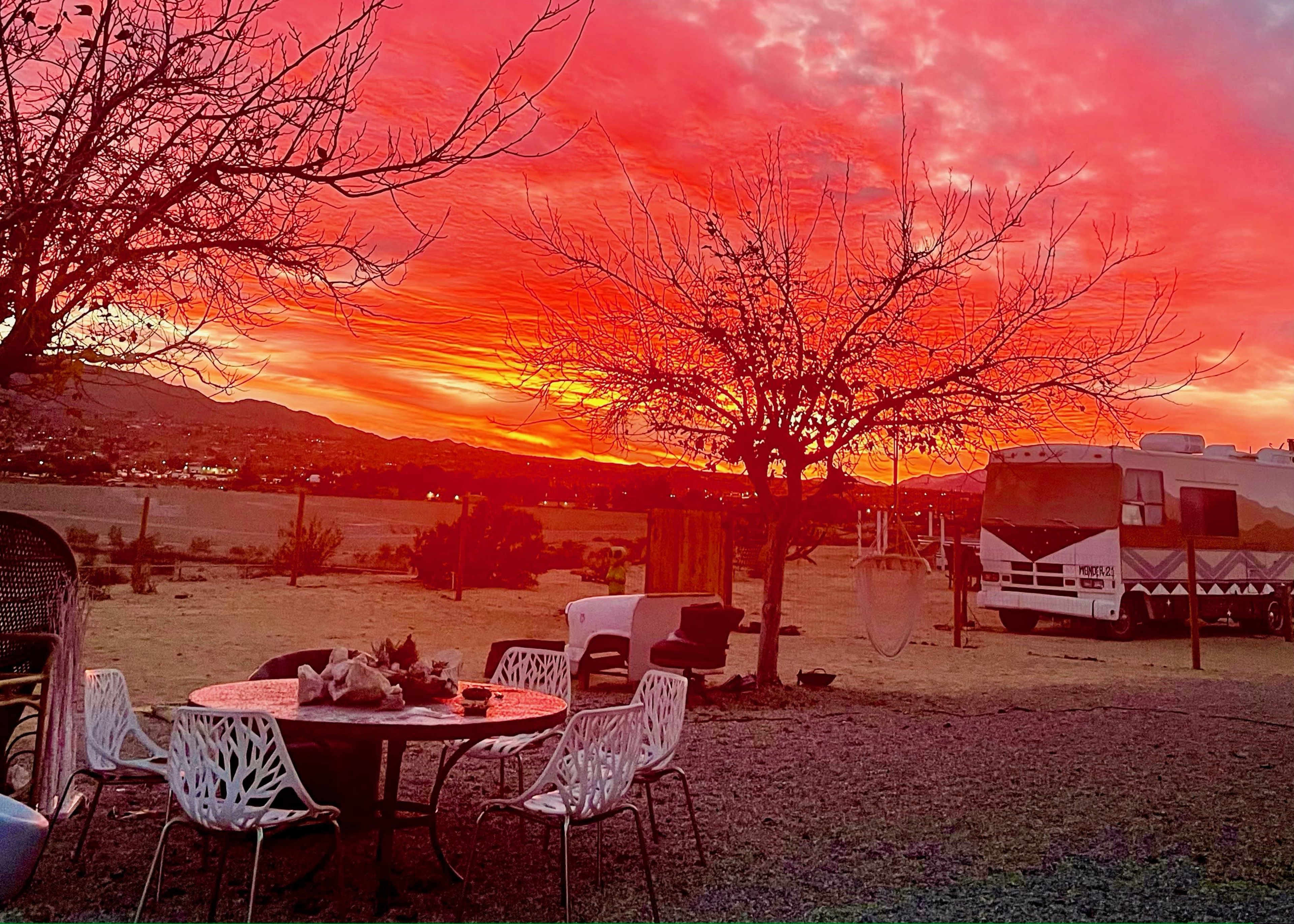 The scene shows a tranquil outdoor setting with a circular table and chairs under a vibrant sunset sky, featuring an RV and barren trees in the background.
