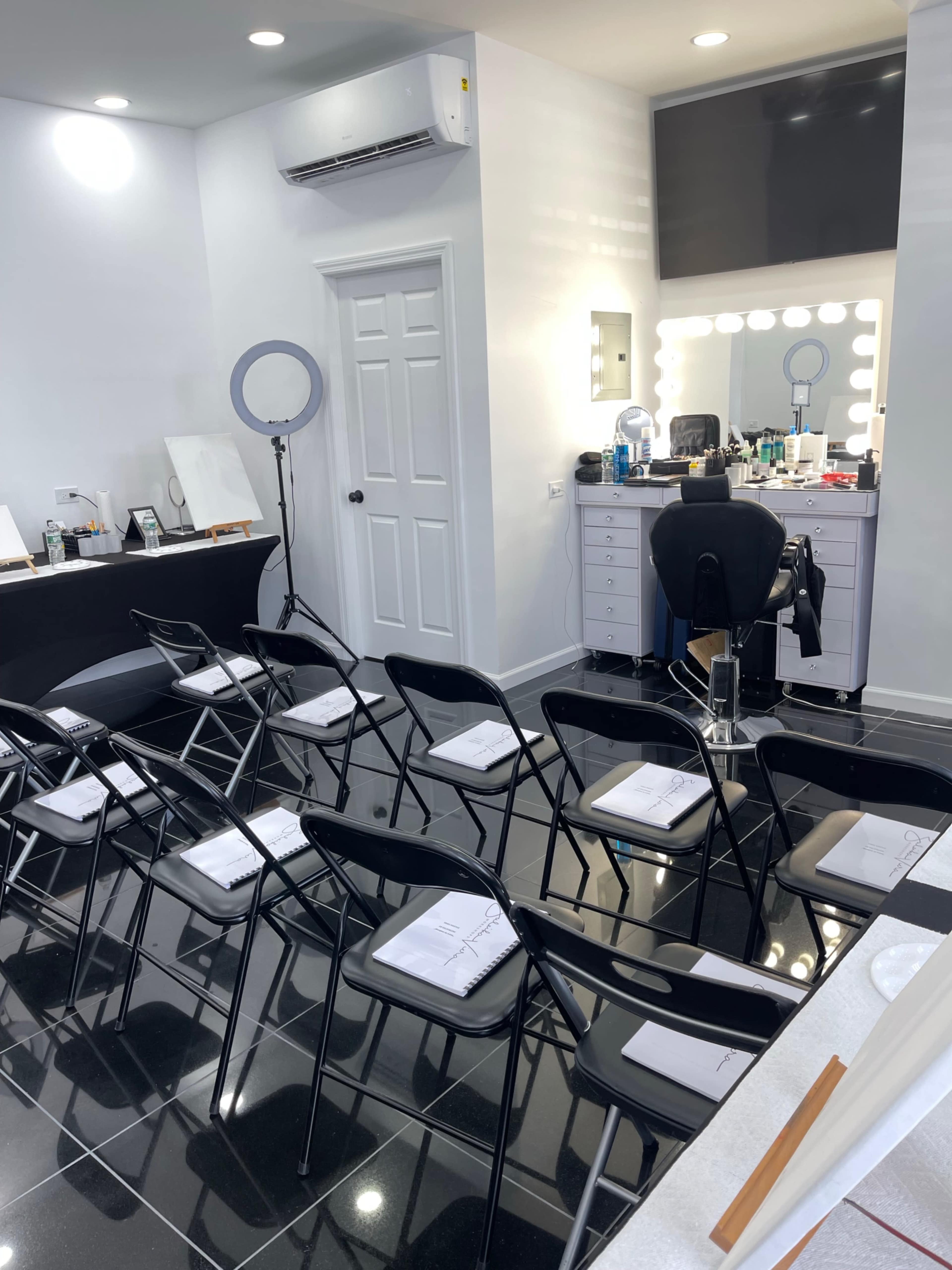 The image shows a beauty studio with rows of black folding chairs facing a makeup station equipped with a mirror and various beauty products.
