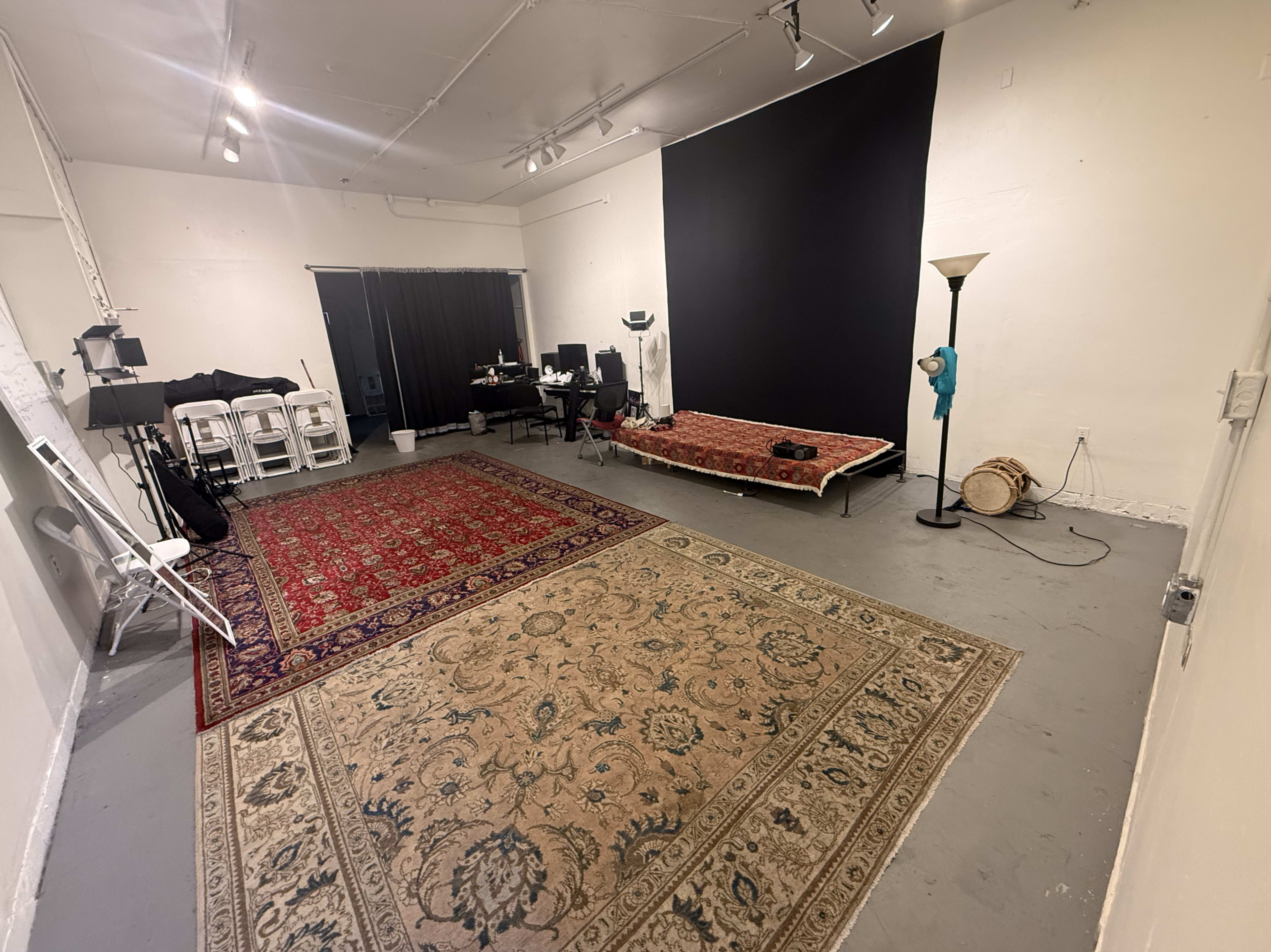 The image shows a spacious room with oriental rugs on the floor, a couch covered with a blanket, a desk with a computer, and a backdrop against one wall.