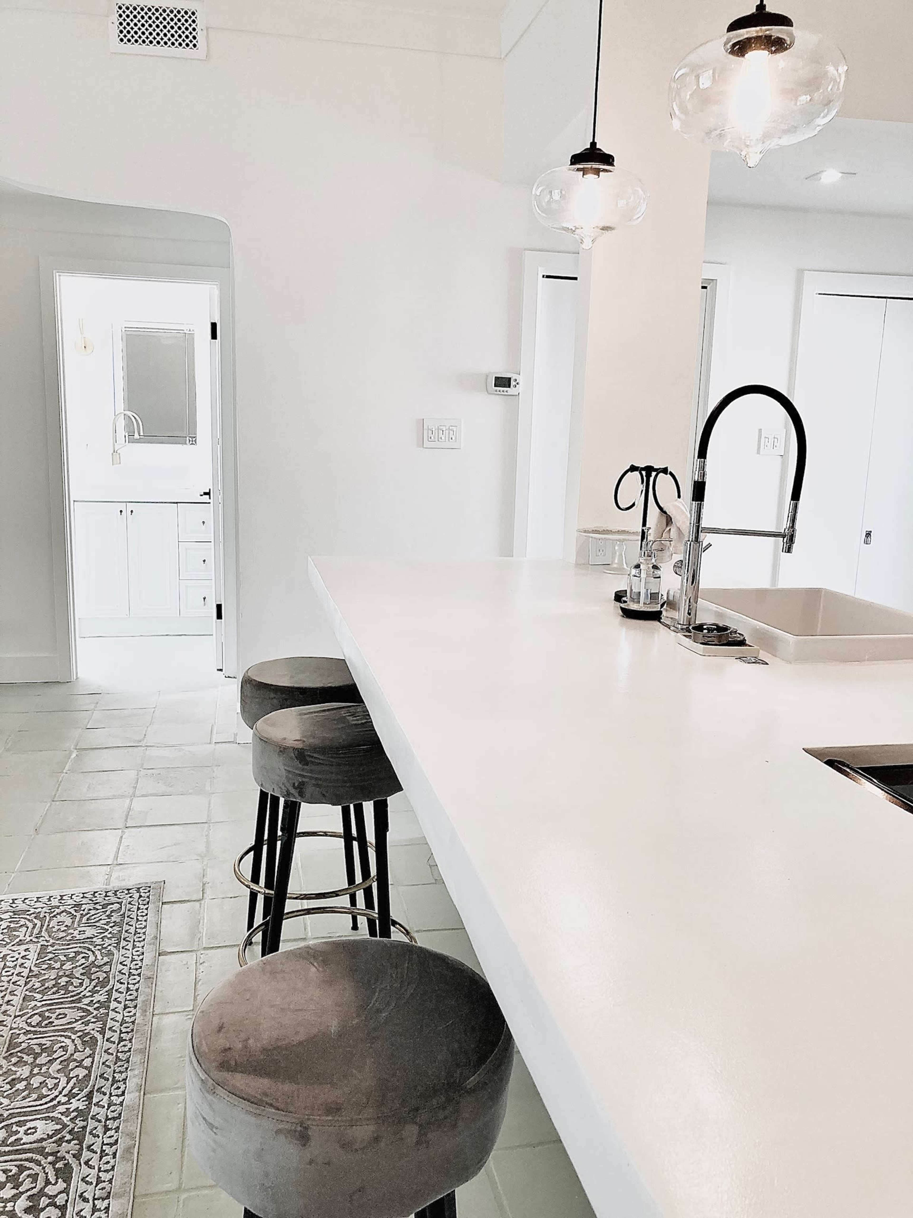 A modern kitchen features a white countertop with three stools and a view into a bathroom through an open door.
