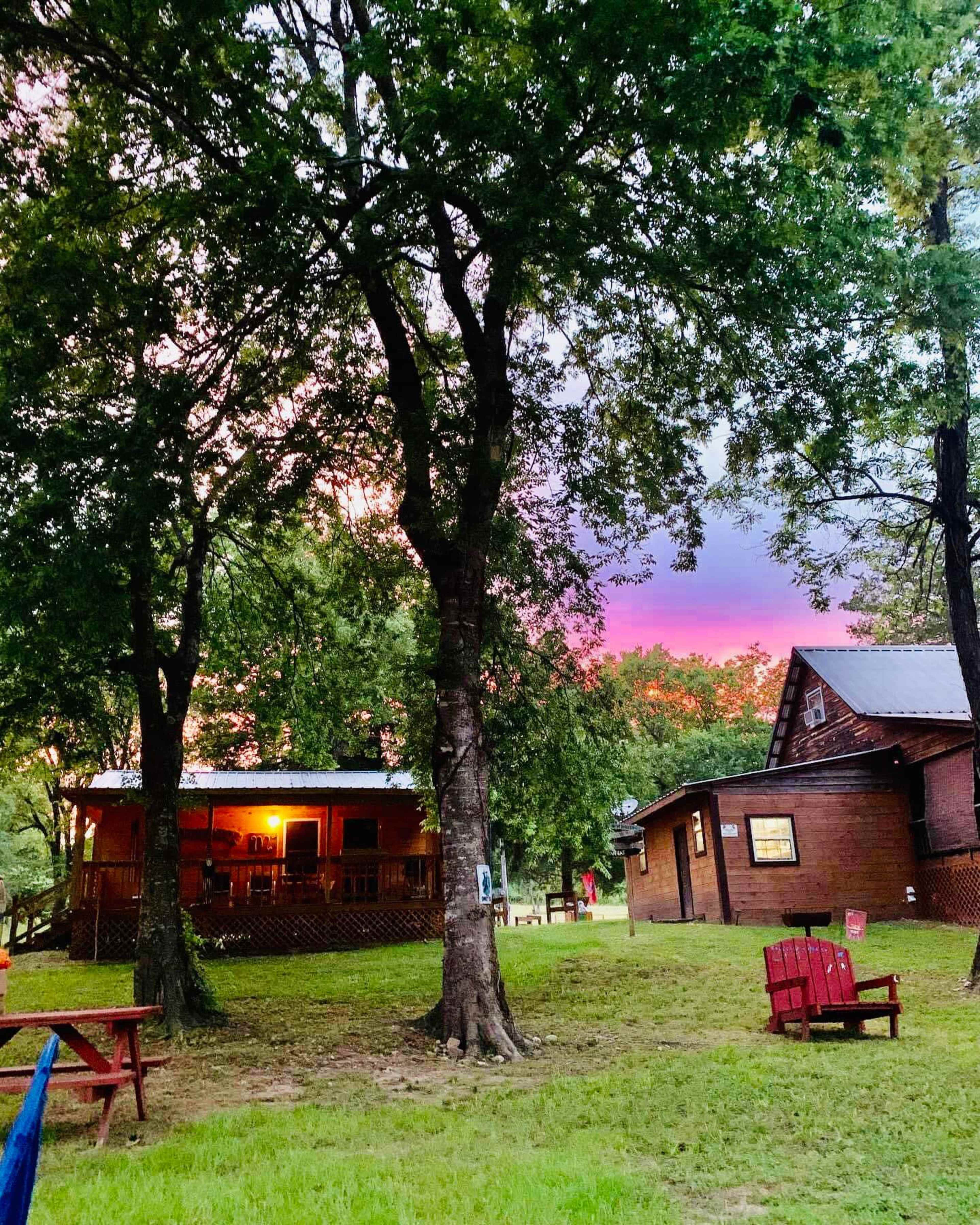 A wooden cabin and an adjacent house are surrounded by trees with a colorful sunset in the background.